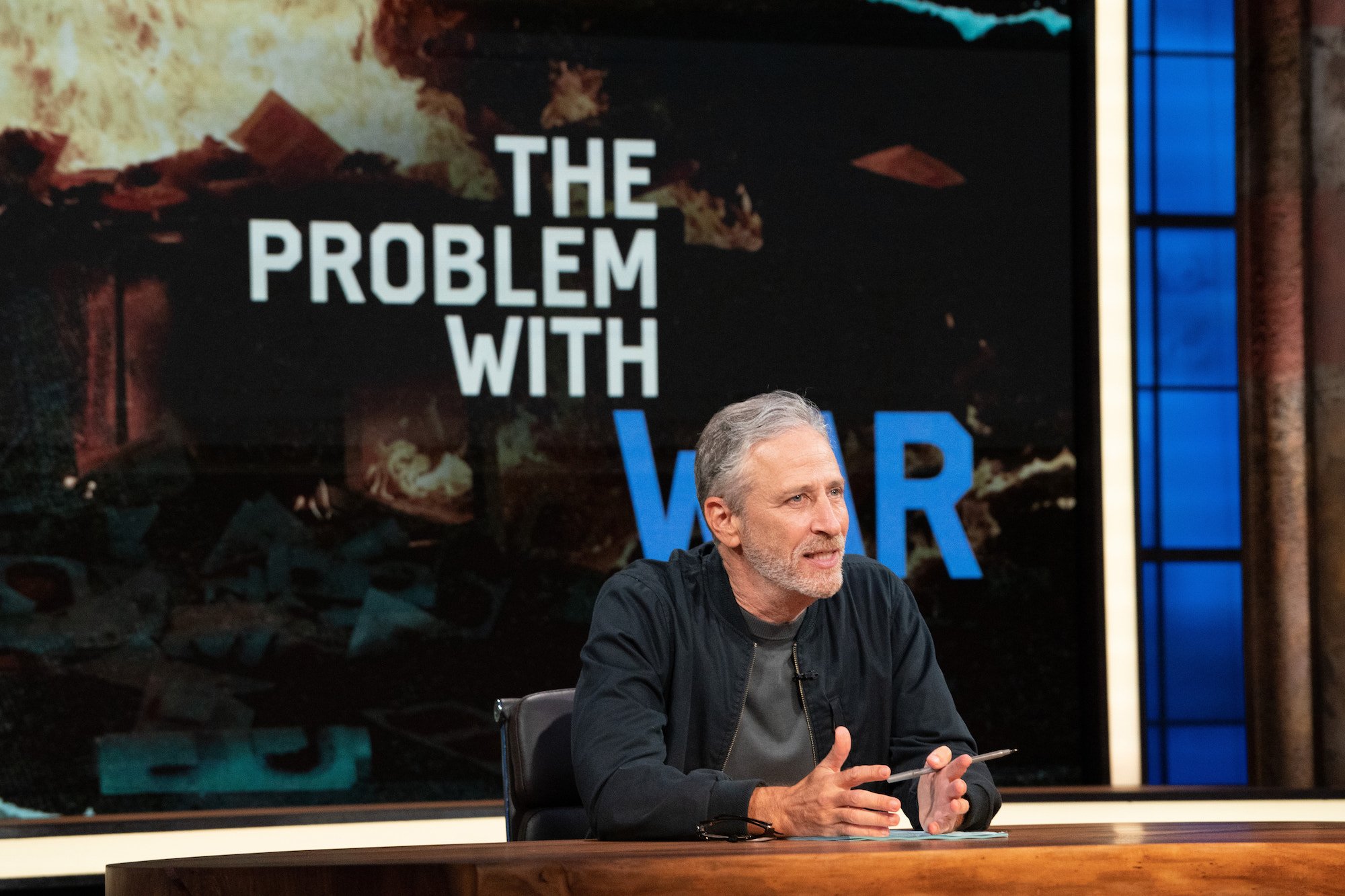 Jon Stewart sits at his desk hosting The Problem