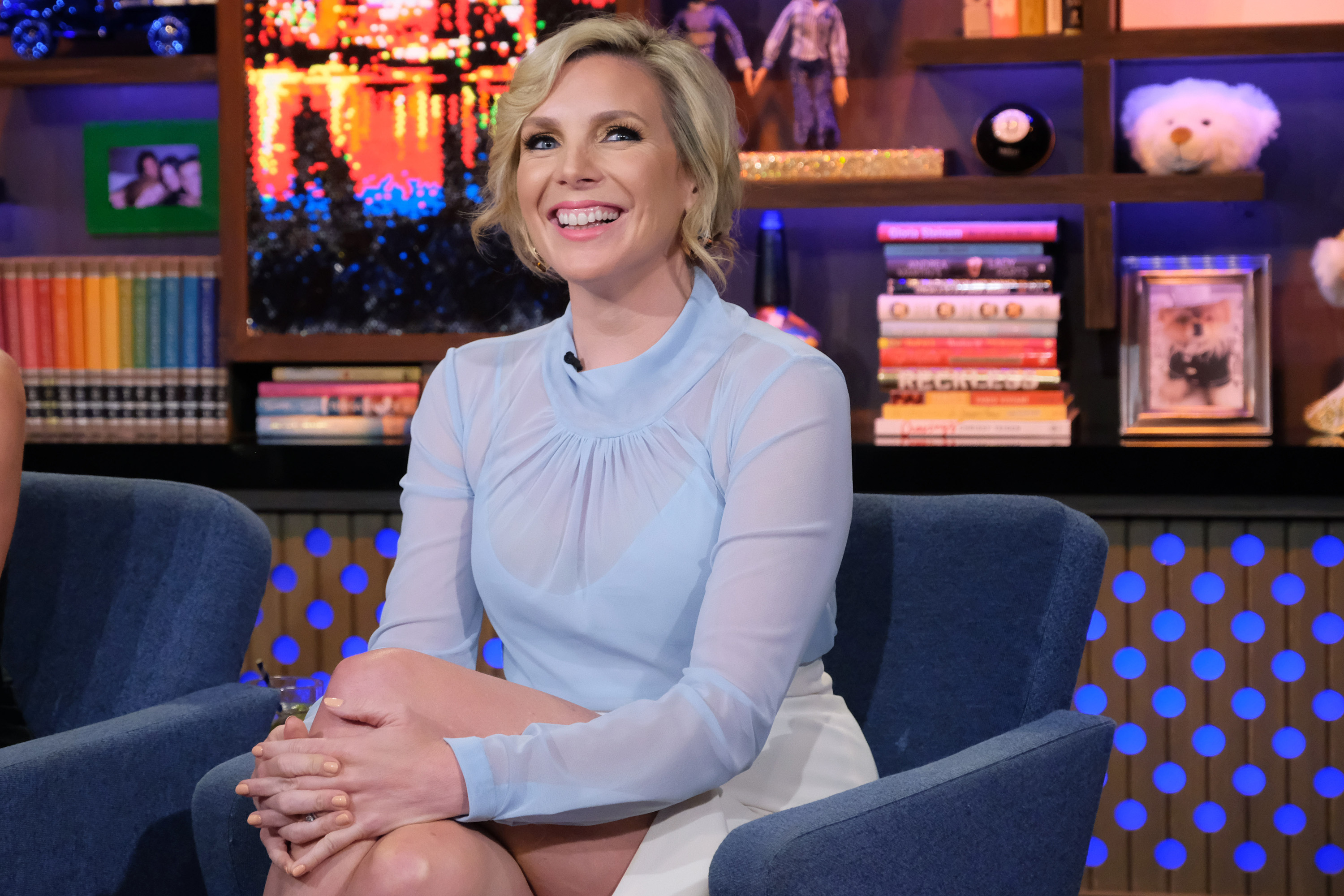 June Diane Raphael sits in a chair and smiles. She is wearing a a long-sleeved blue see-through top and a white skirt.