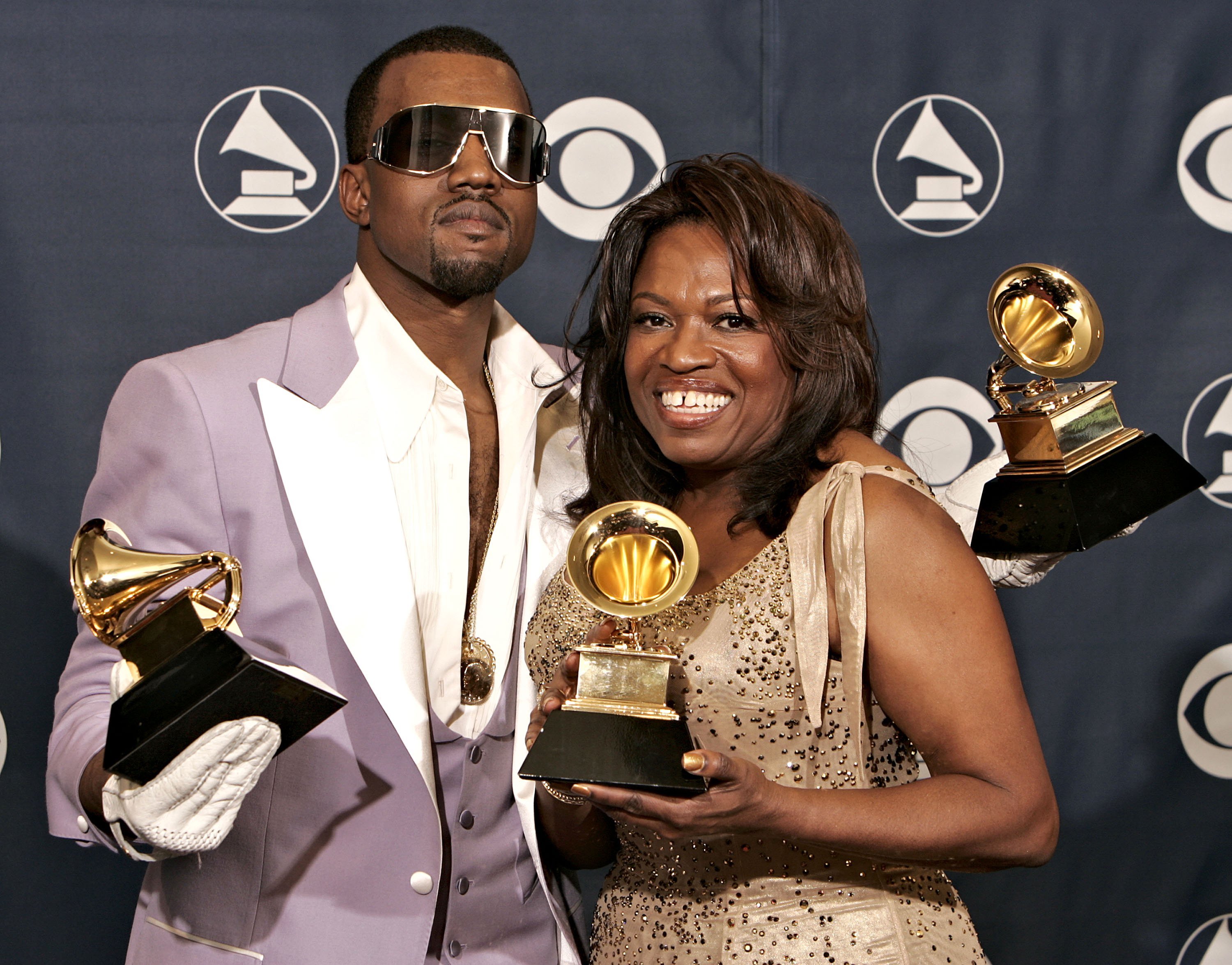 Kanye West and his mom, Donda, holding his three Grammy Awards while smiling for the camera on the red carpet.