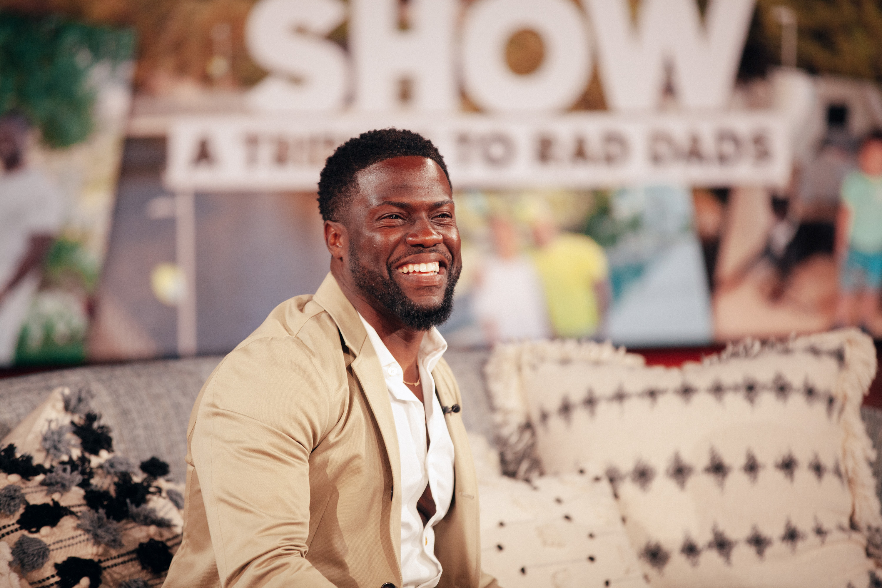 Kevin Hart sits on a couch wearing a tan suit.