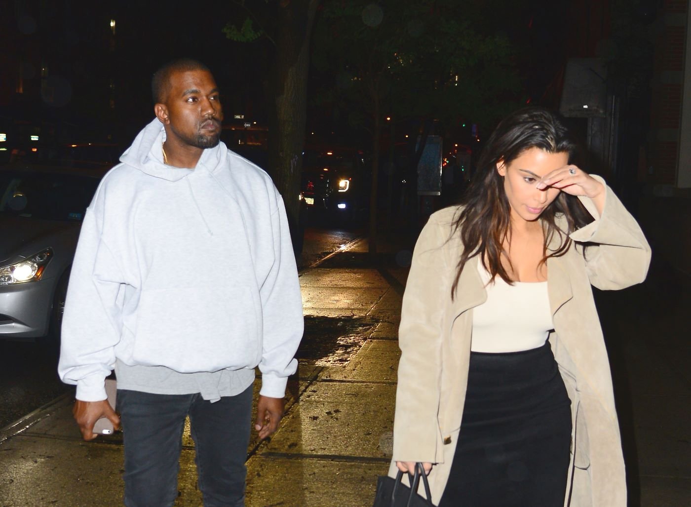 Kim Kardashian West and Kanye West walking down a dark street. Kanye is wearing a white hoodie and blue jean pants. Kim is wearing a black pair of pants, white shirt, and brown trench coat. 