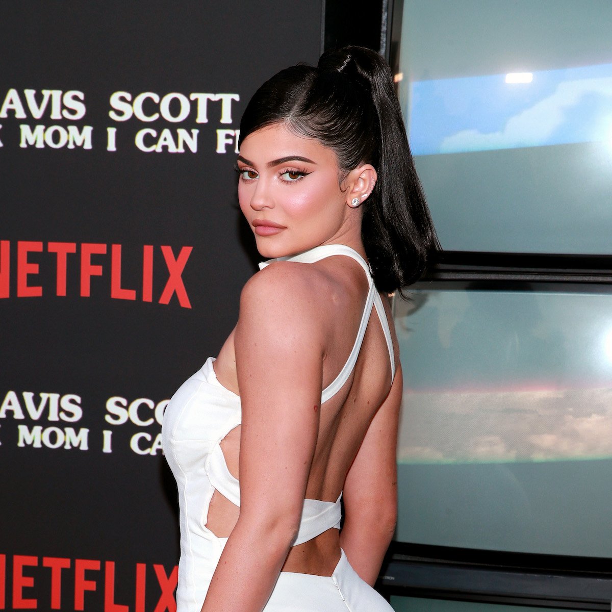 Kylie Jenner looks over her shoulder at the camera wearing a white dress and a high ponytail.