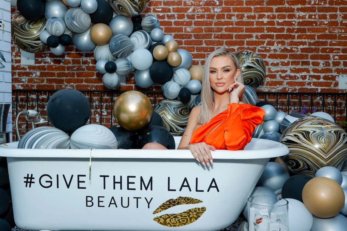 Lala Kent wears an orange dress and poses in a bathtub emblazoned with "Give Them Lala Beauty."