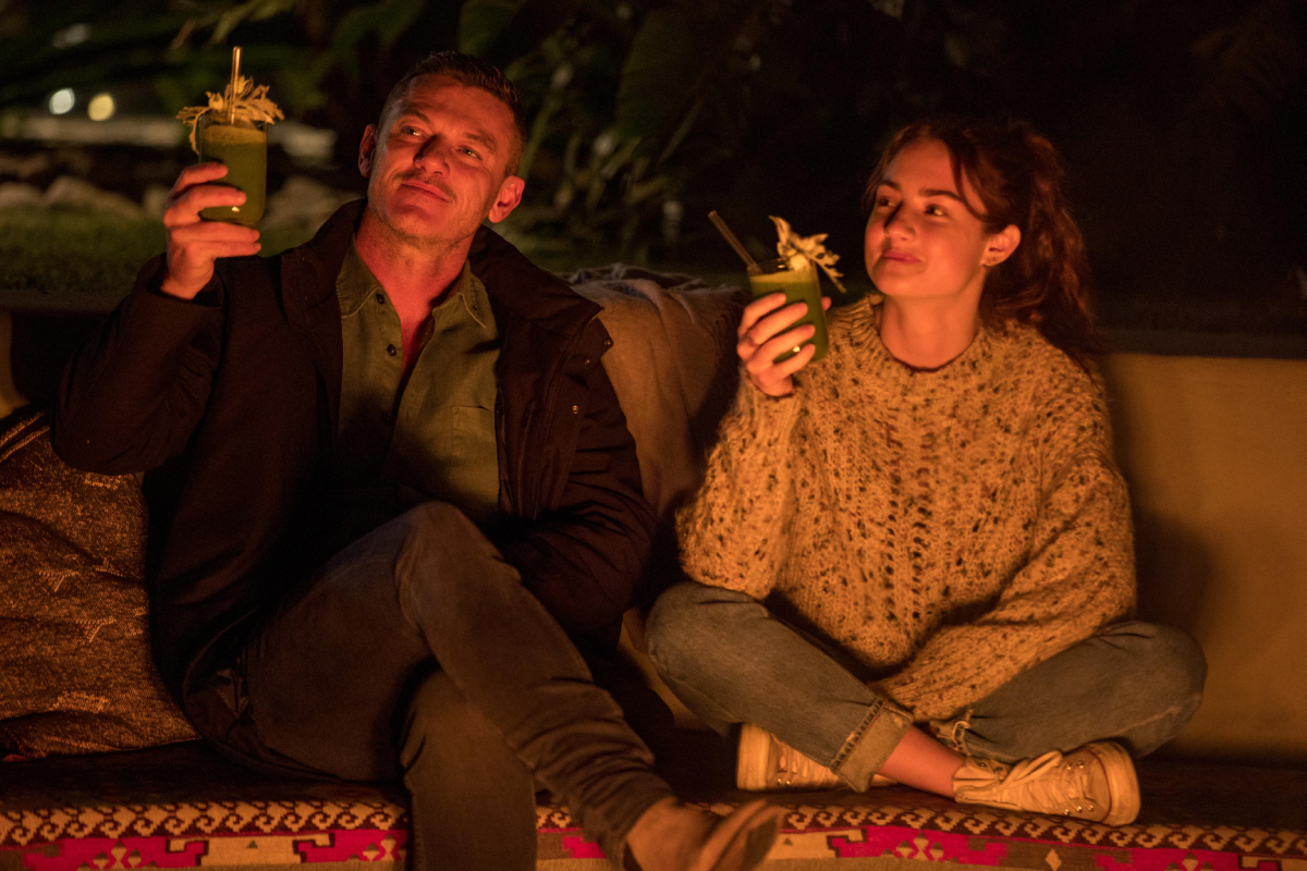 Lars and Zoe raise their smoothie glasses for a cheers. Zoe wears a sweater and jeans and Lars wears pants, a collared shirt, and a jacket.