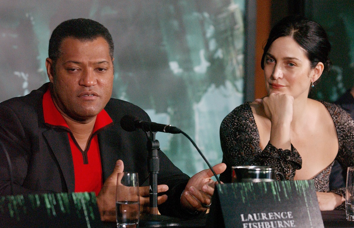 Carrie-Anne Moss with her hand on her chin looks at Laurence Fishburne as he speaks into a microphone at an event for ‘The Matrix.’