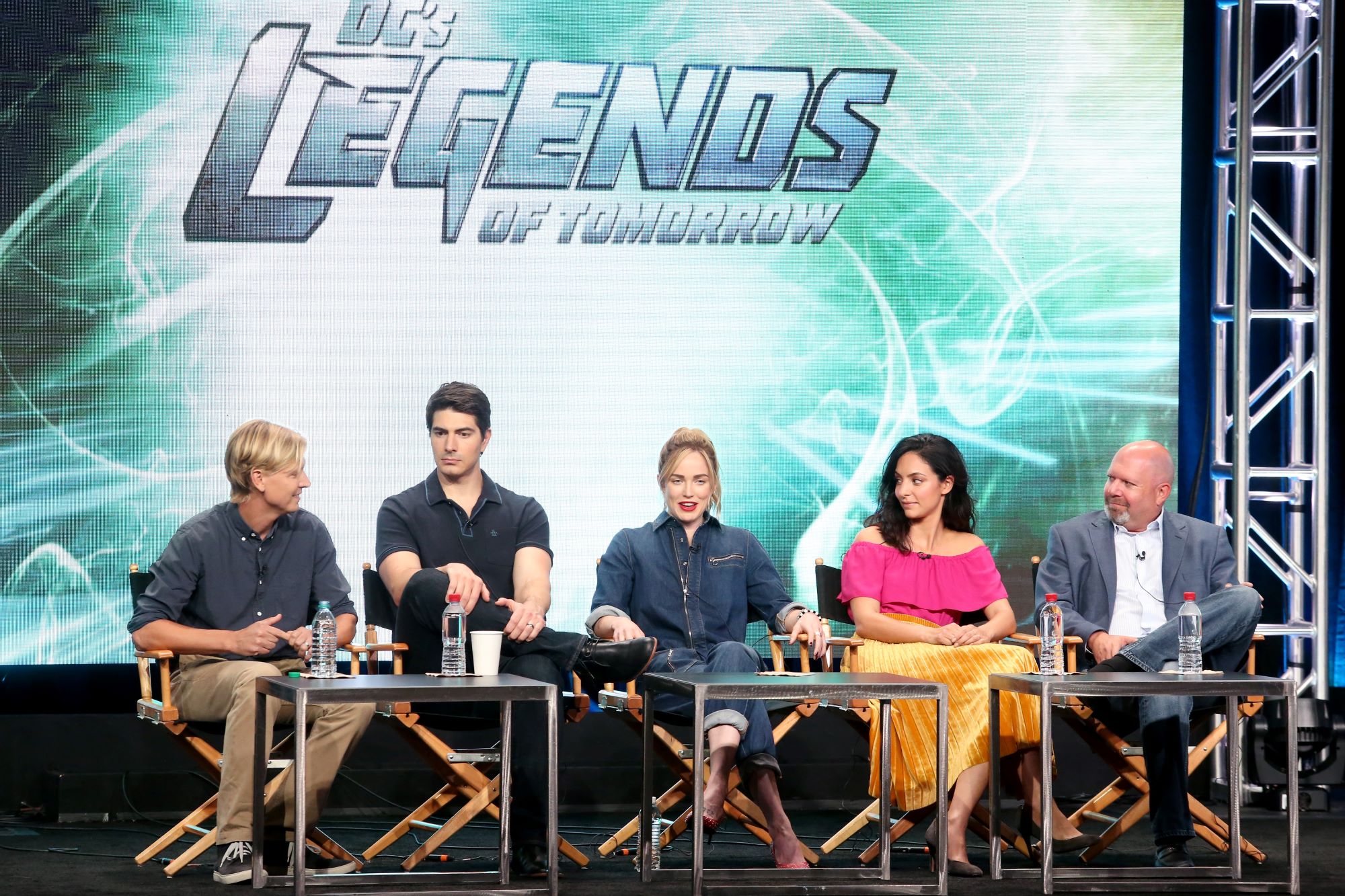 Phil Klemmer, Brandon Routh, Caity Lotz, Tala Ashe, and Marc Guggenheim of 'DC's Legends of Tomorrow' speak onstage about the show. They sit in director's chairs with the 'DC's Legends of Tomorrow' title card on the screen behind them. 'DC's Legends of Tomorrow' Season 7 Episode 1 premieres on Oct. 13.