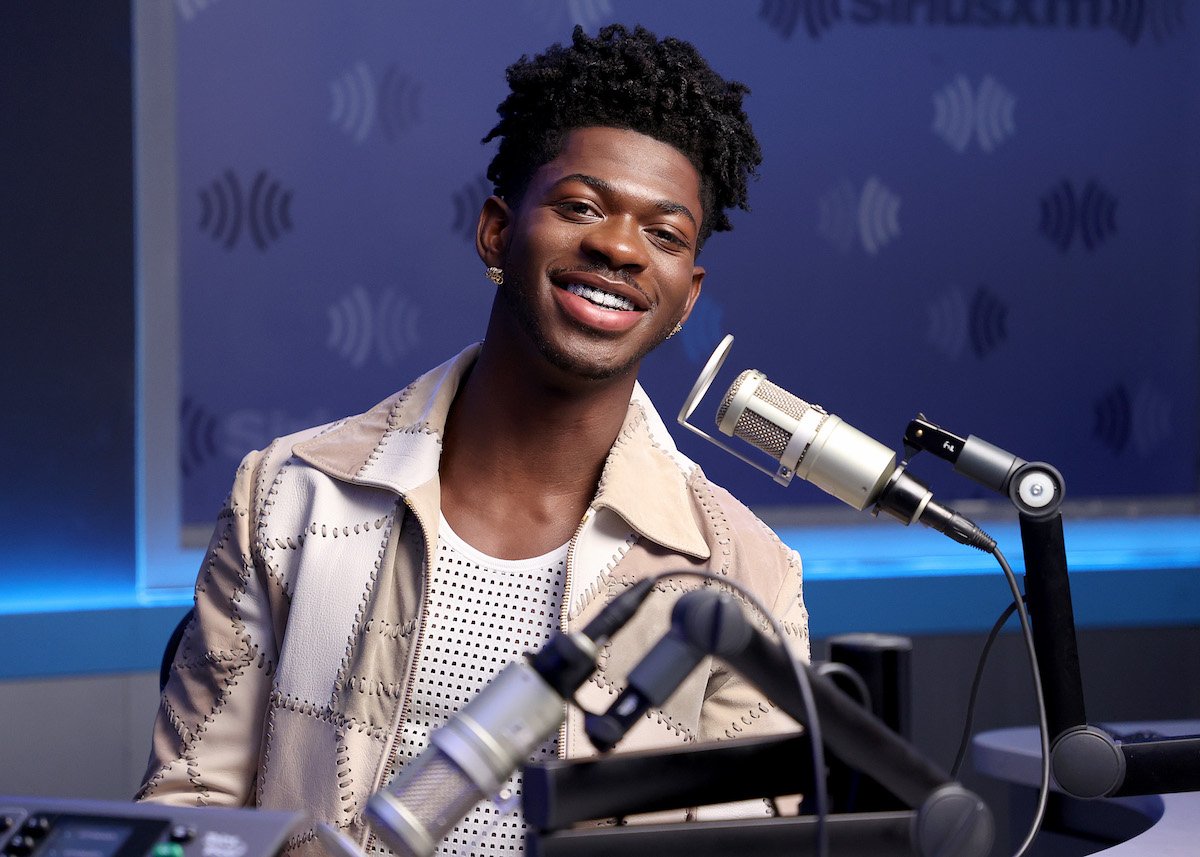 Lil Nas X dressed casually smiles and looks at the camera with a microphone in front of him during an interview.
