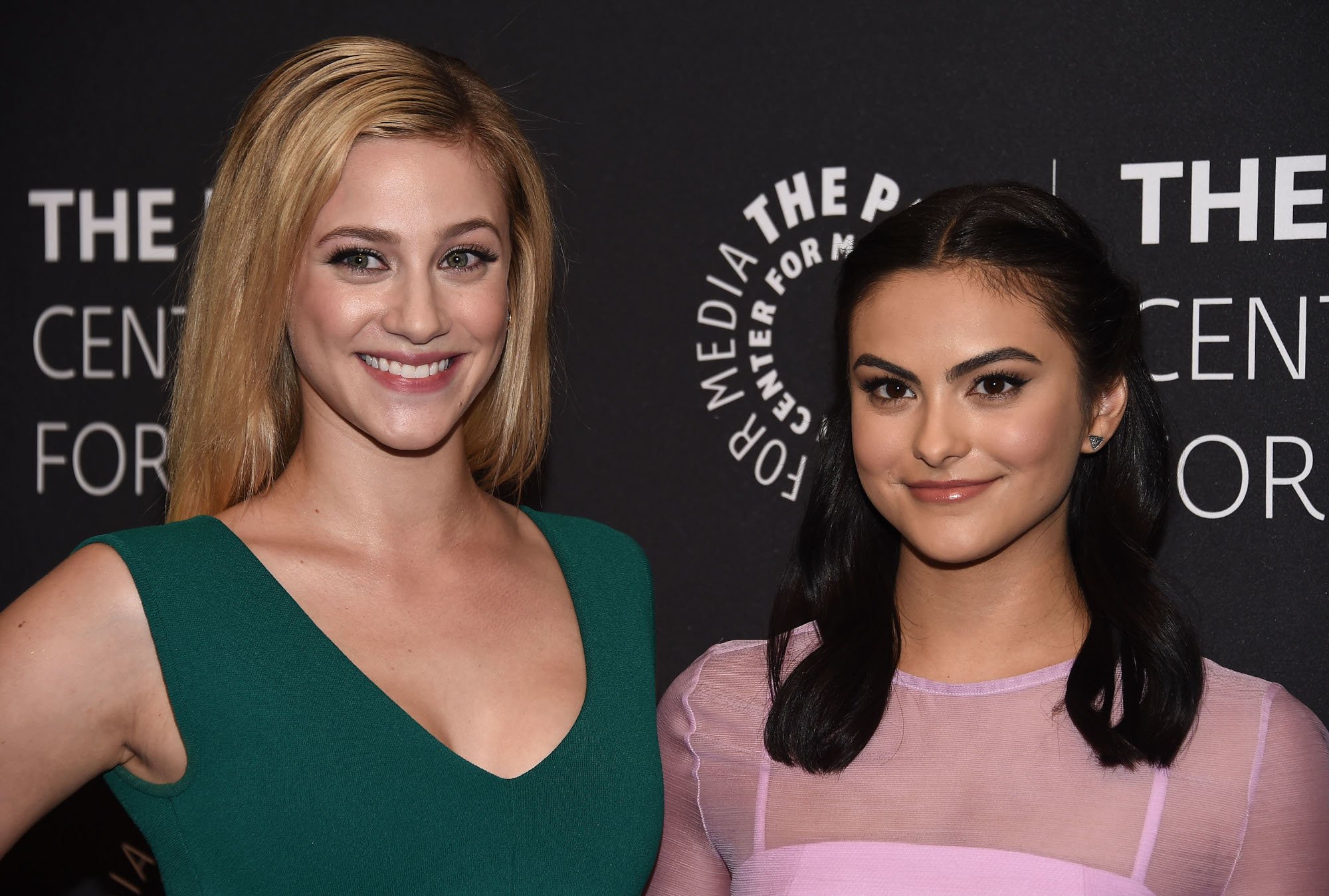 'Riverdale' Season 5 stars Lili Reinhart and Camila Mendes. They're standing side by side. Reinhart is wearing a green v-neck dress with her blonde hair down. Mendes is wearing a pink dress with her black hair half-up and half-down.
