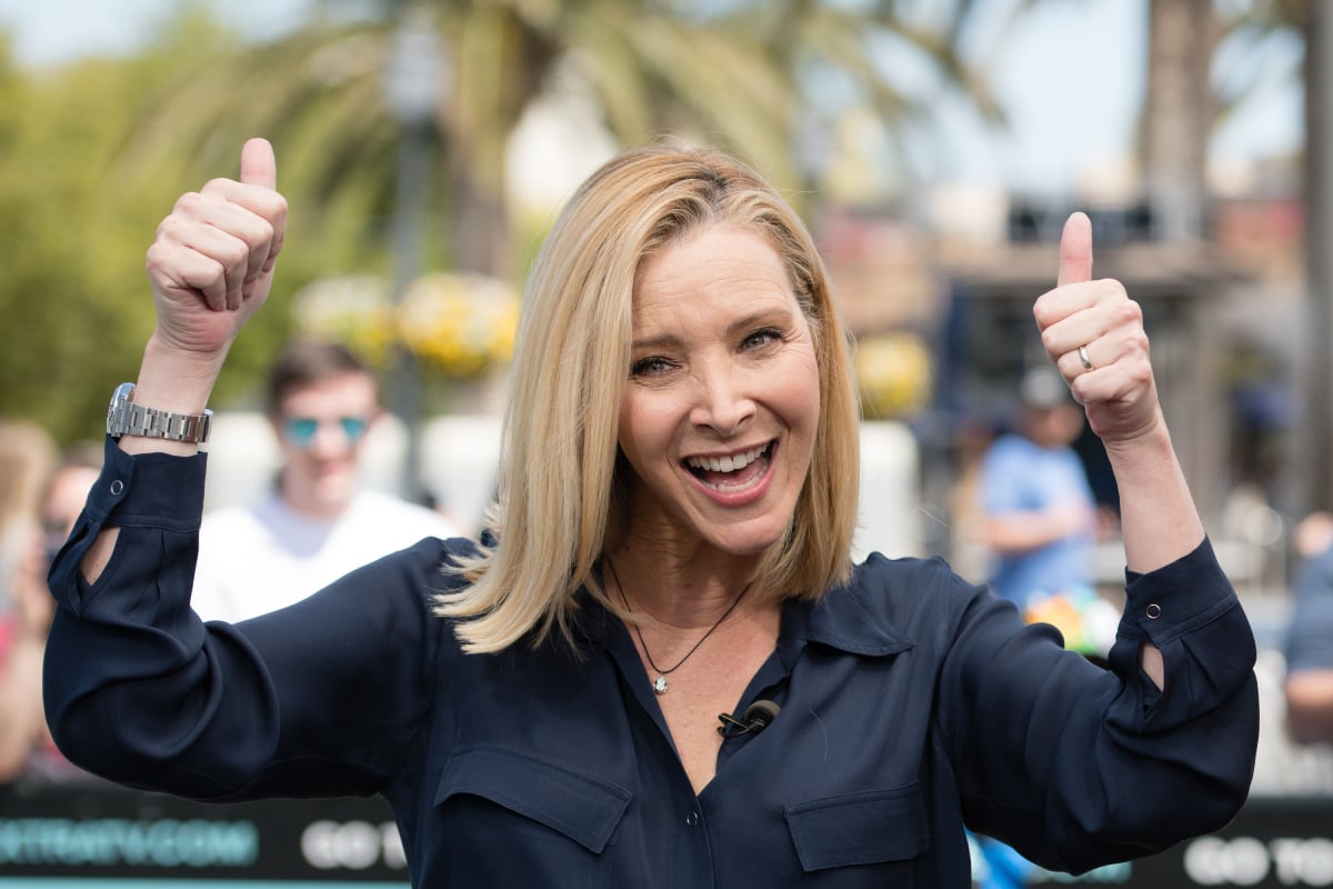 Lisa Kudrow gives two thumbs up and smiles. She is wearing a navy blue button-down shirt.