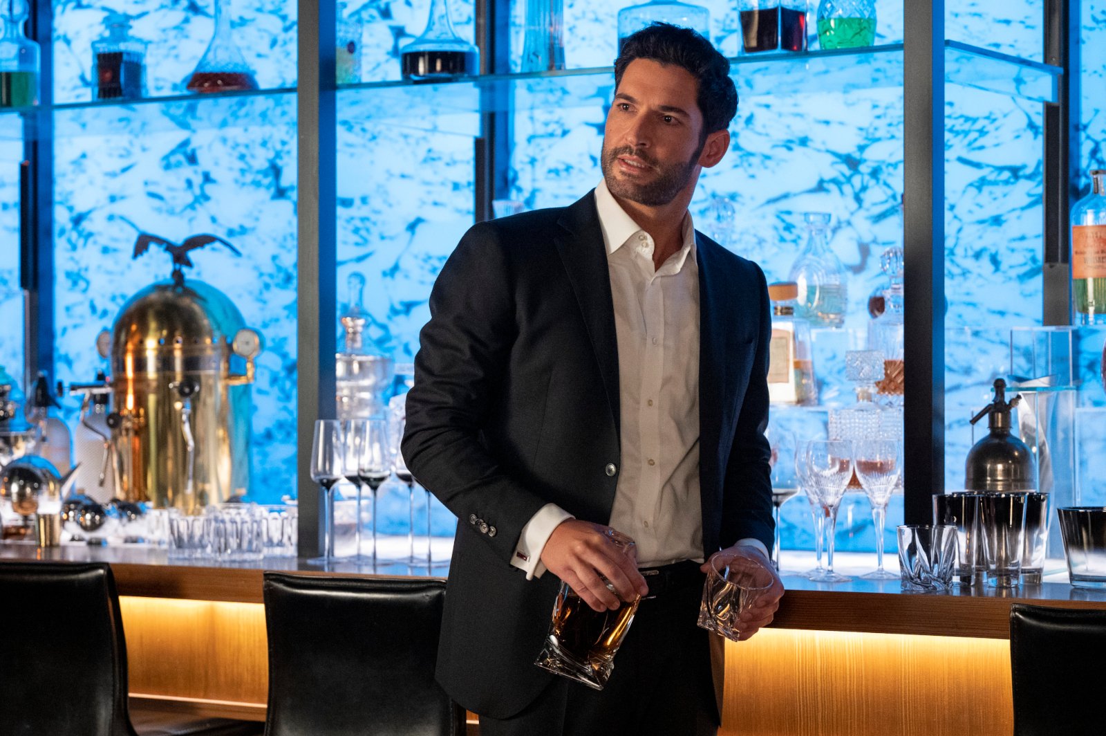 Tom Ellis as Lucifer Morningstar in 'Lucifer' Season 6. He's wearing a suit and standing in front of a bar. He's holding a glass and a bottle. 'Lucifer' will not receive a season 7.