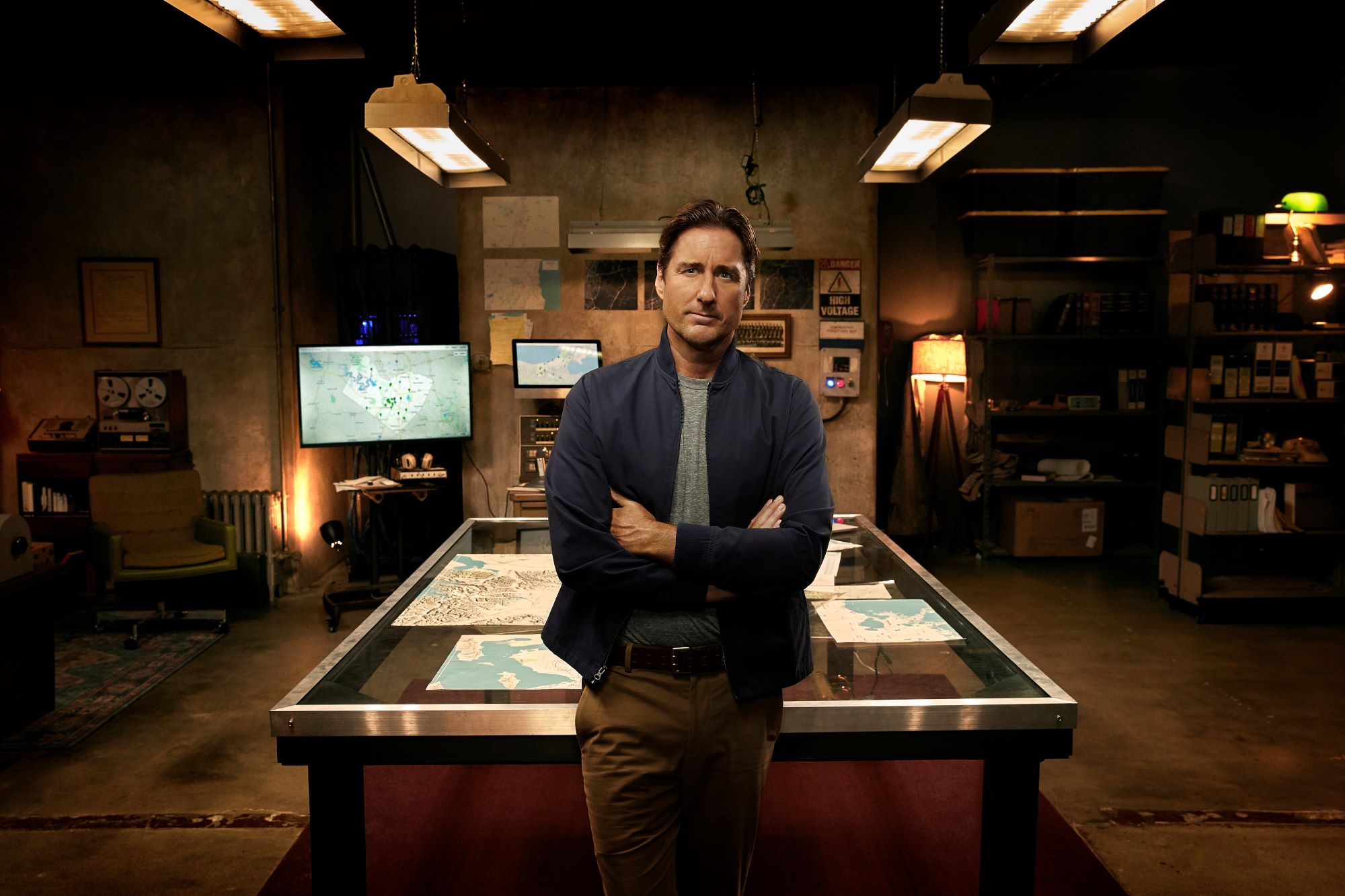 'Stargirl' Season 3 actor Luke Wilson wears tan pants, a gray shirt, and a dark blue jacket as he leans against a glass table.