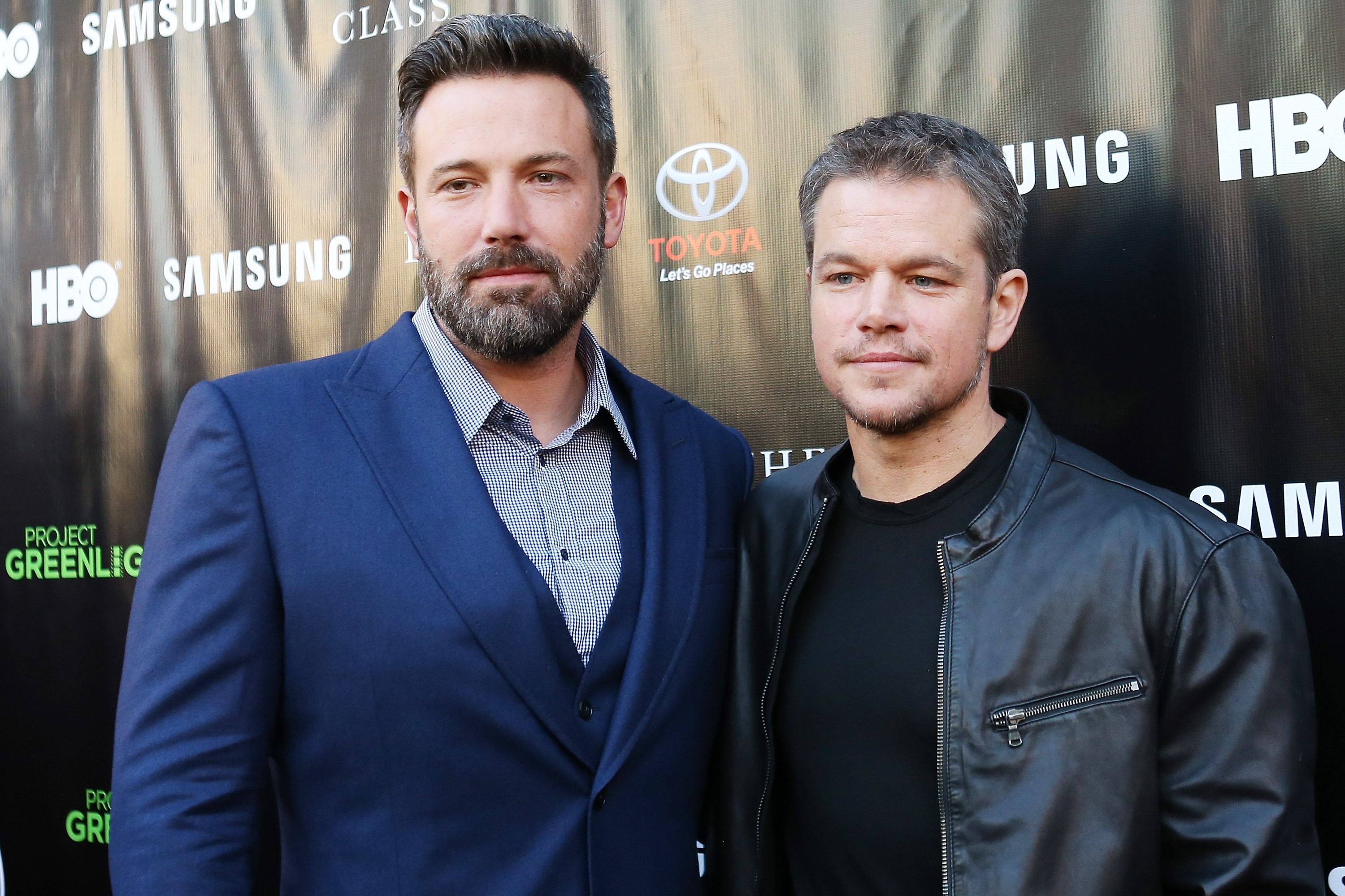 Ben Affleck, in a blue suit with a blue shirt, and Matt Damon, in a black t-shirt and black leather jacket, at an event in 2015.