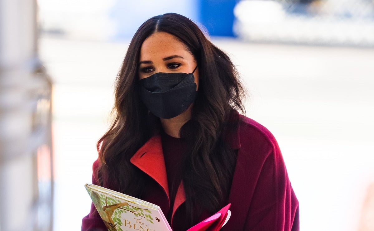 Meghan Markle holding her children's book during a visit with schoolkids in Harlem, wearing a red coat and a black mask