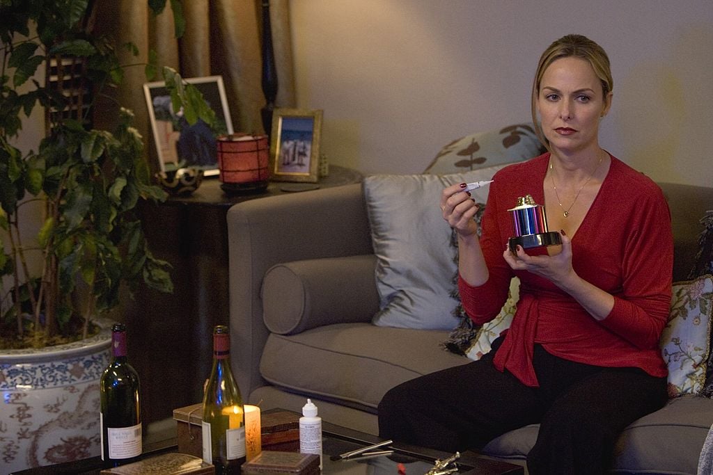 Melora Hardin as Jan Levinson sits on the couch in a red shirt.
