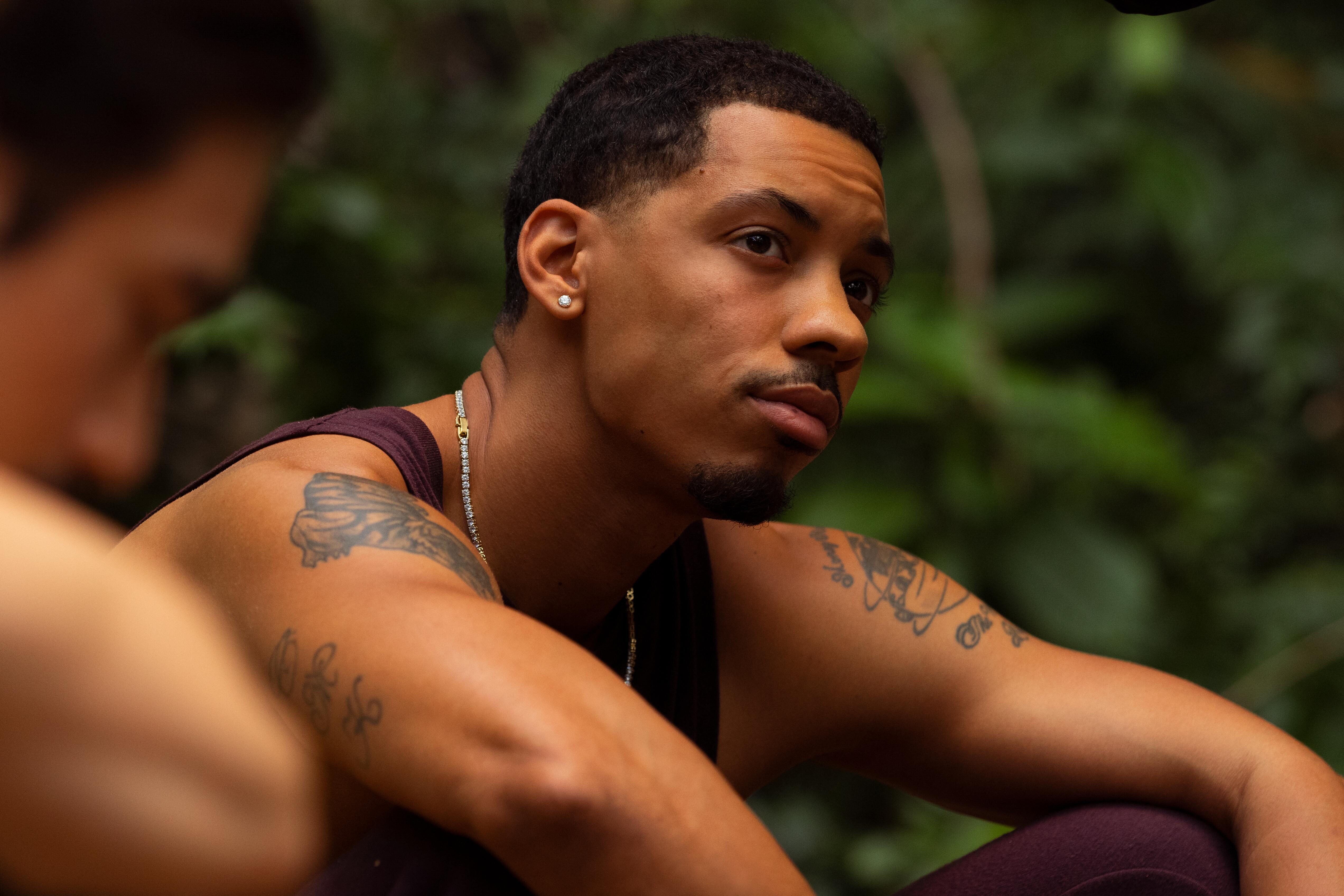 Ben Chandler sits on the ground and looks up. He is wearing stud earrings, a maroon tank top, and a silver chain.