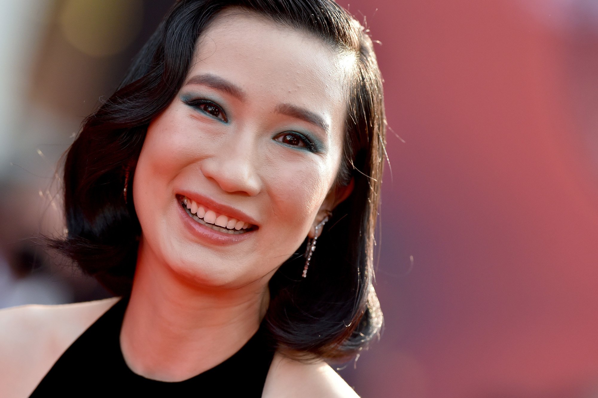 'Shang-Chi' star Meng'er Zhang on the red carpet for the Marvel film. She's smiling and has green eye makeup. You can see the top of her dress, which is black and has tanktop sleeves. The background behind her is blurred but includes a lot of red.