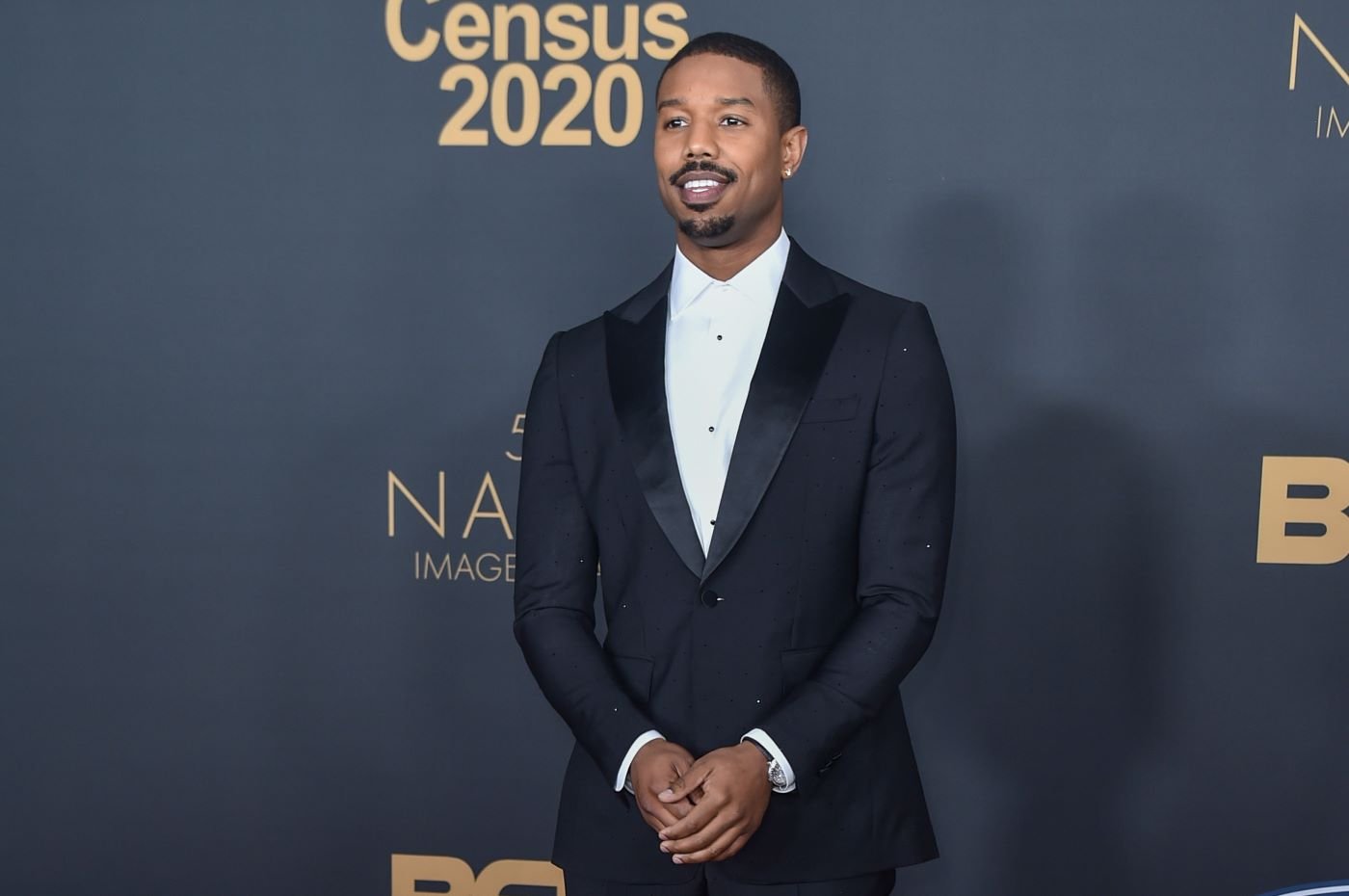 Michael B. Jordan dressed in a black suit against a grey background.