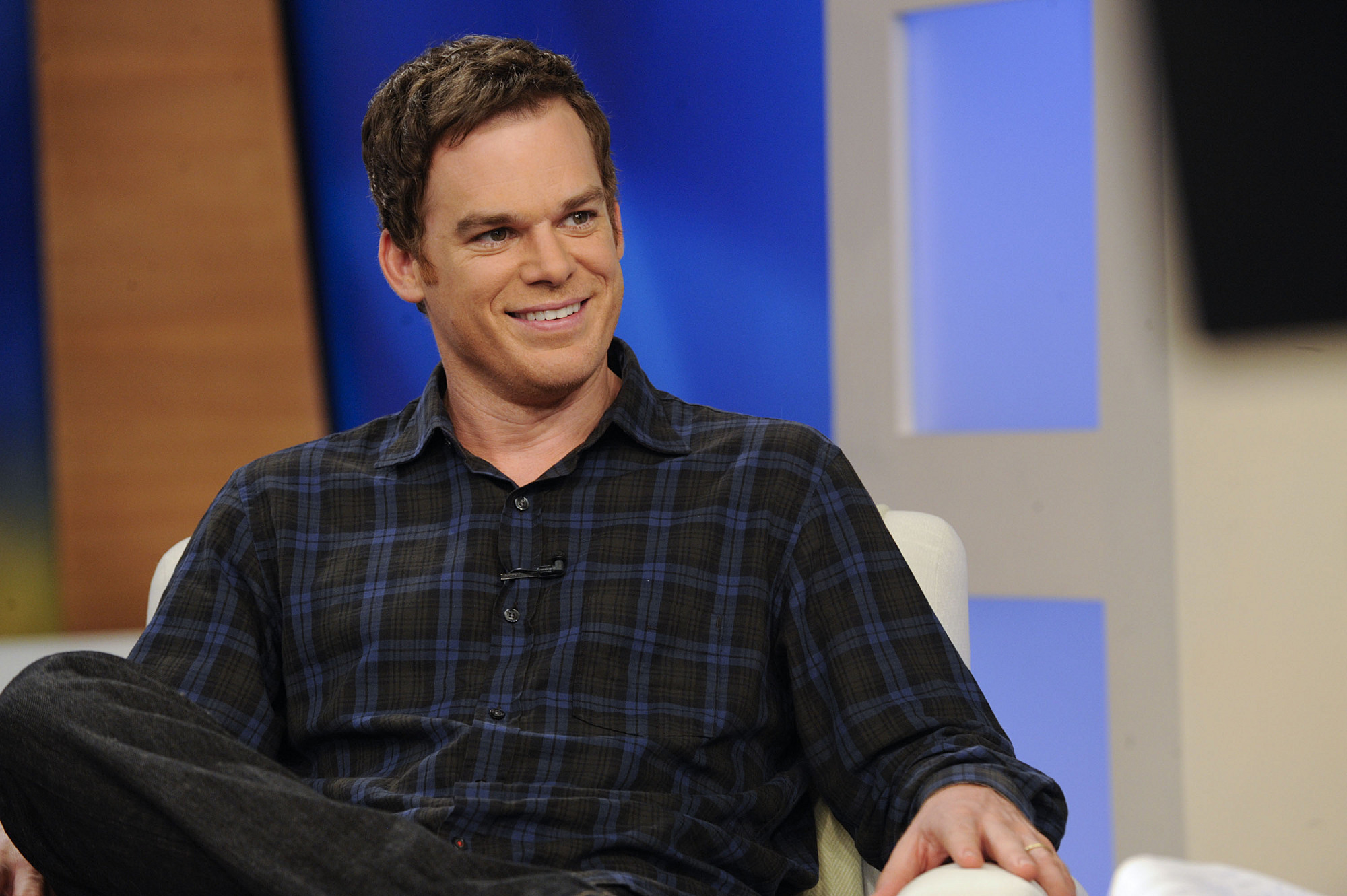 Michael C. Hall sits in a chair with his legs crossed wearing a black and blue plaid shirt.