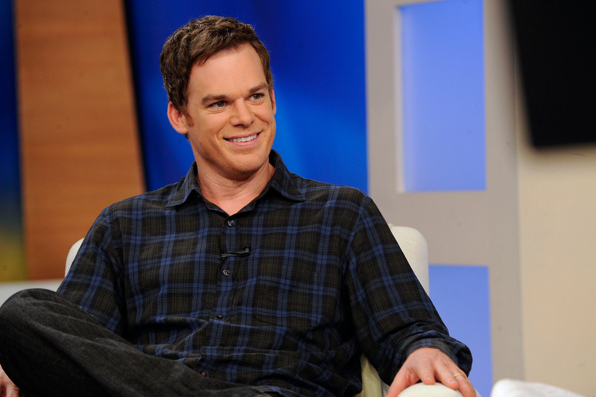 Michael C. Hall sits in a chair with his legs crossed wearing a black and blue plaid shirt.