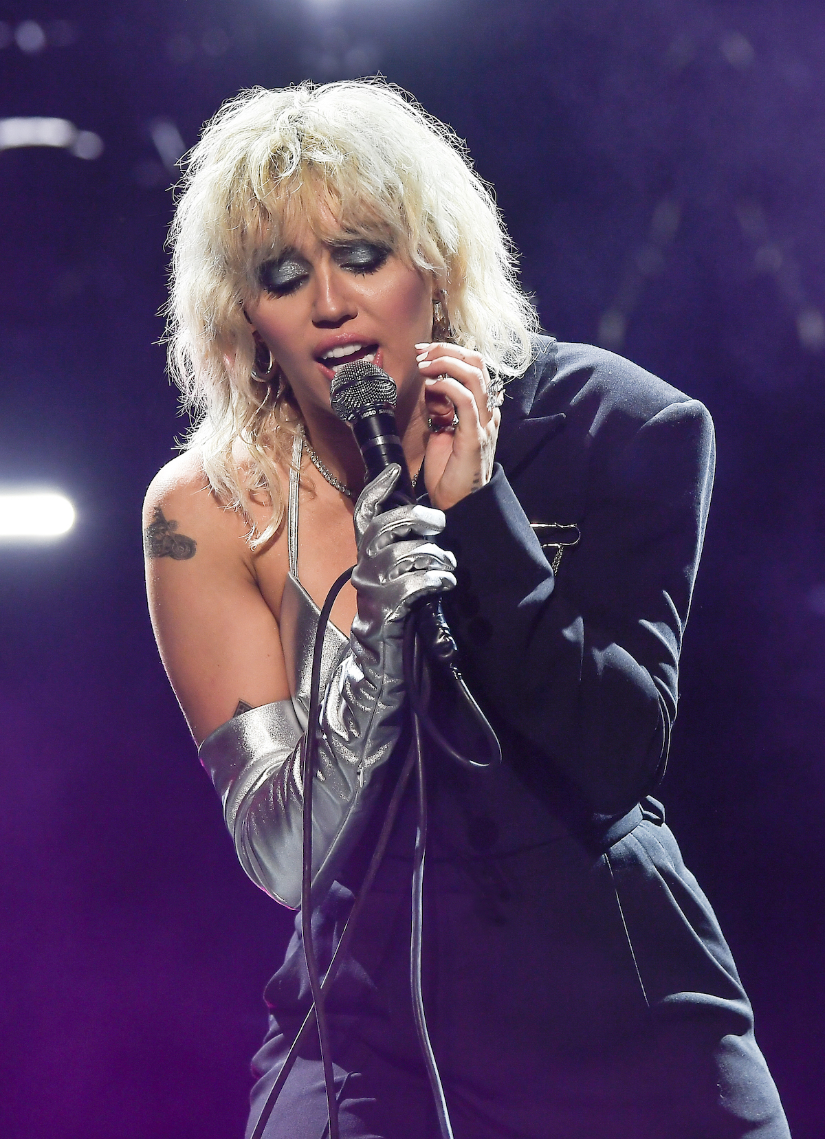 Miley Cyrus wearing silver eyeshadow sings passionately into a microphone on stage.
