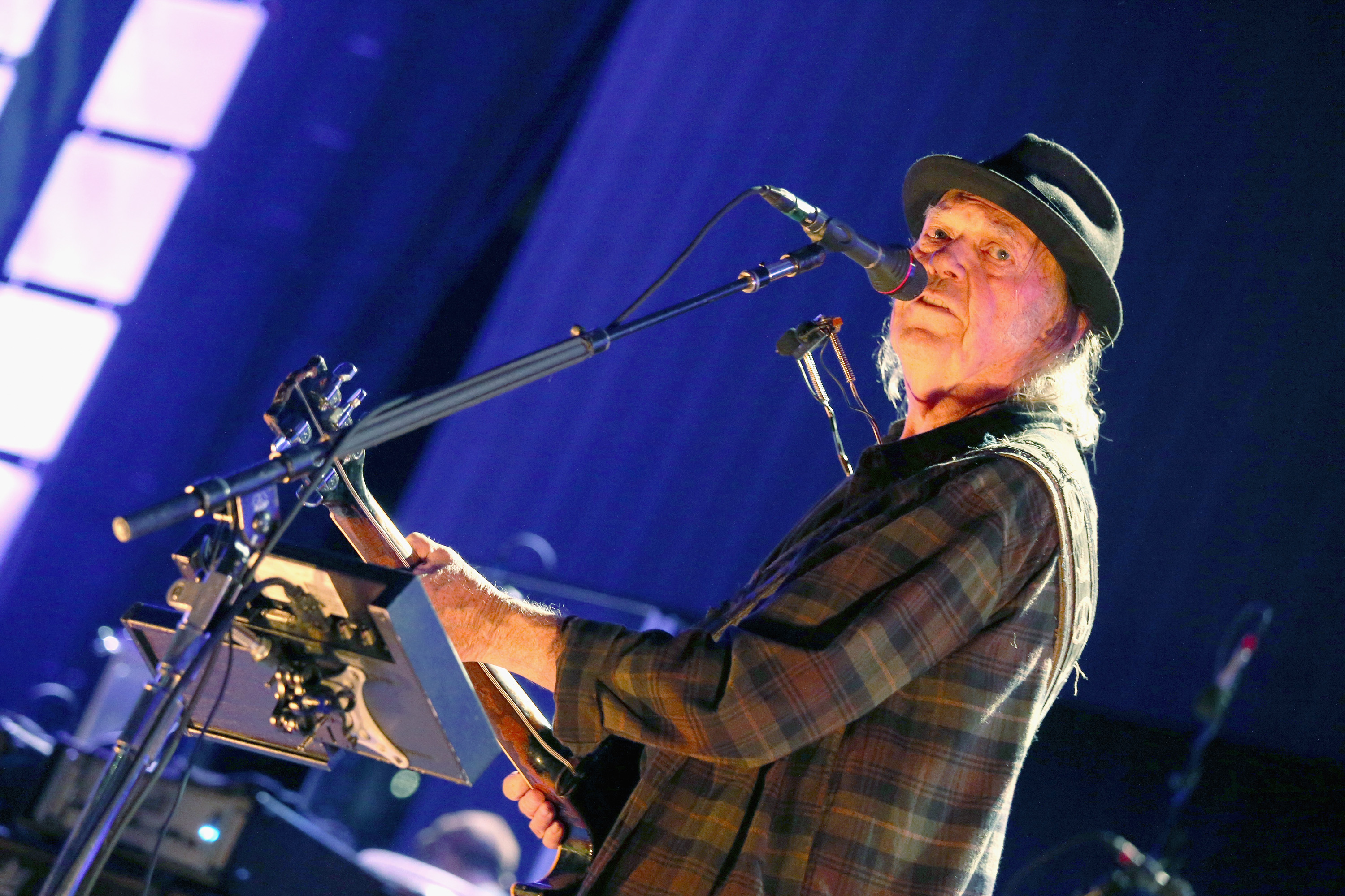 Neil Young performs on stage in a hat and flannel shirt.