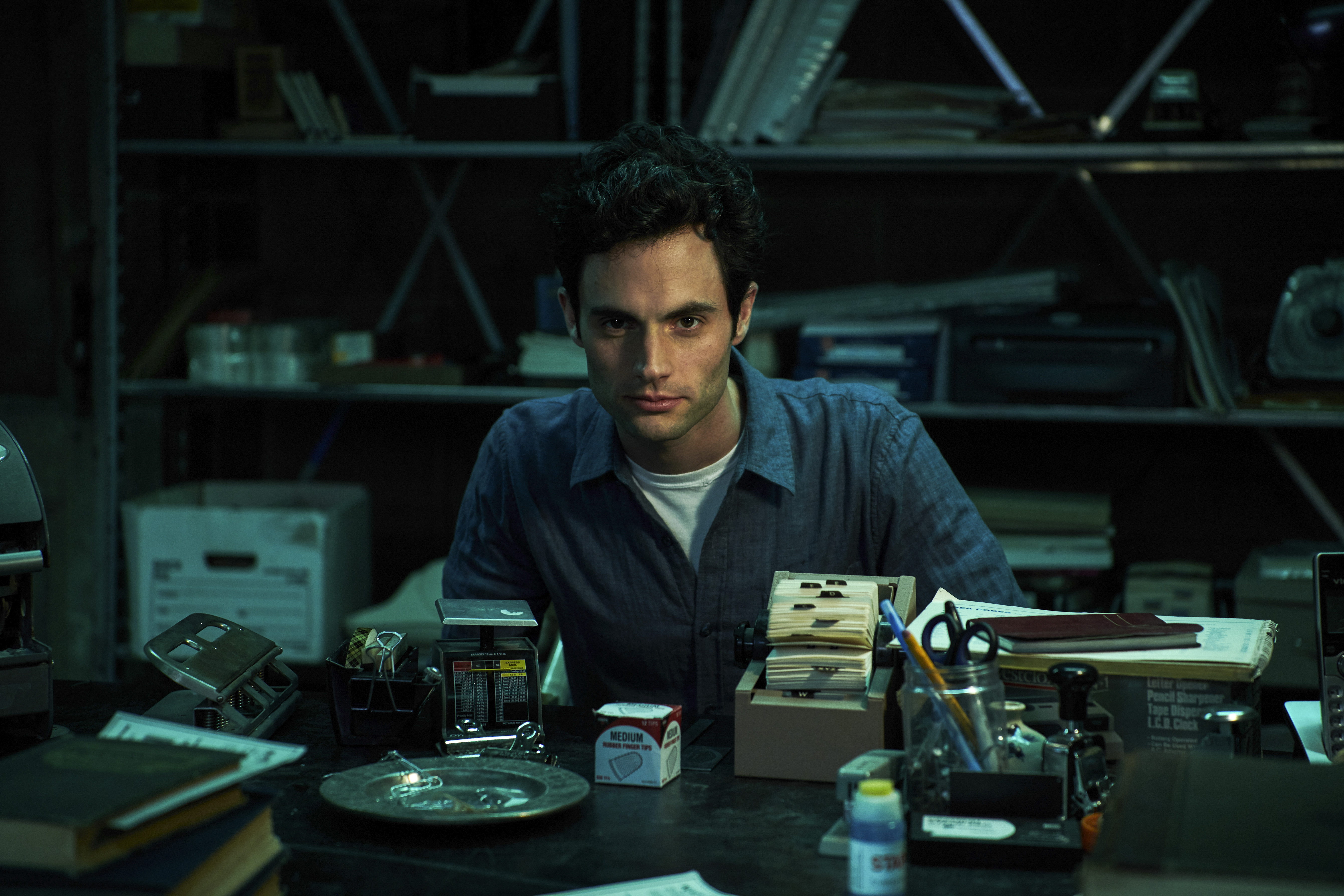 Joe Goldberg sits behind a desk and looks menacingly forward. He is wearing a blue button-down shirt.