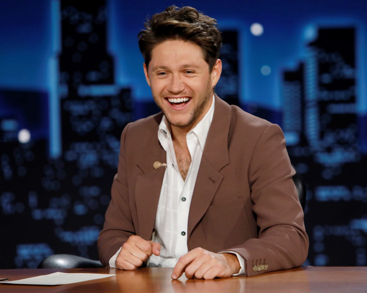Niall Horan dressed in a white dress shirt and a black suit jacket sitting at a desk in front of a black and blue background.