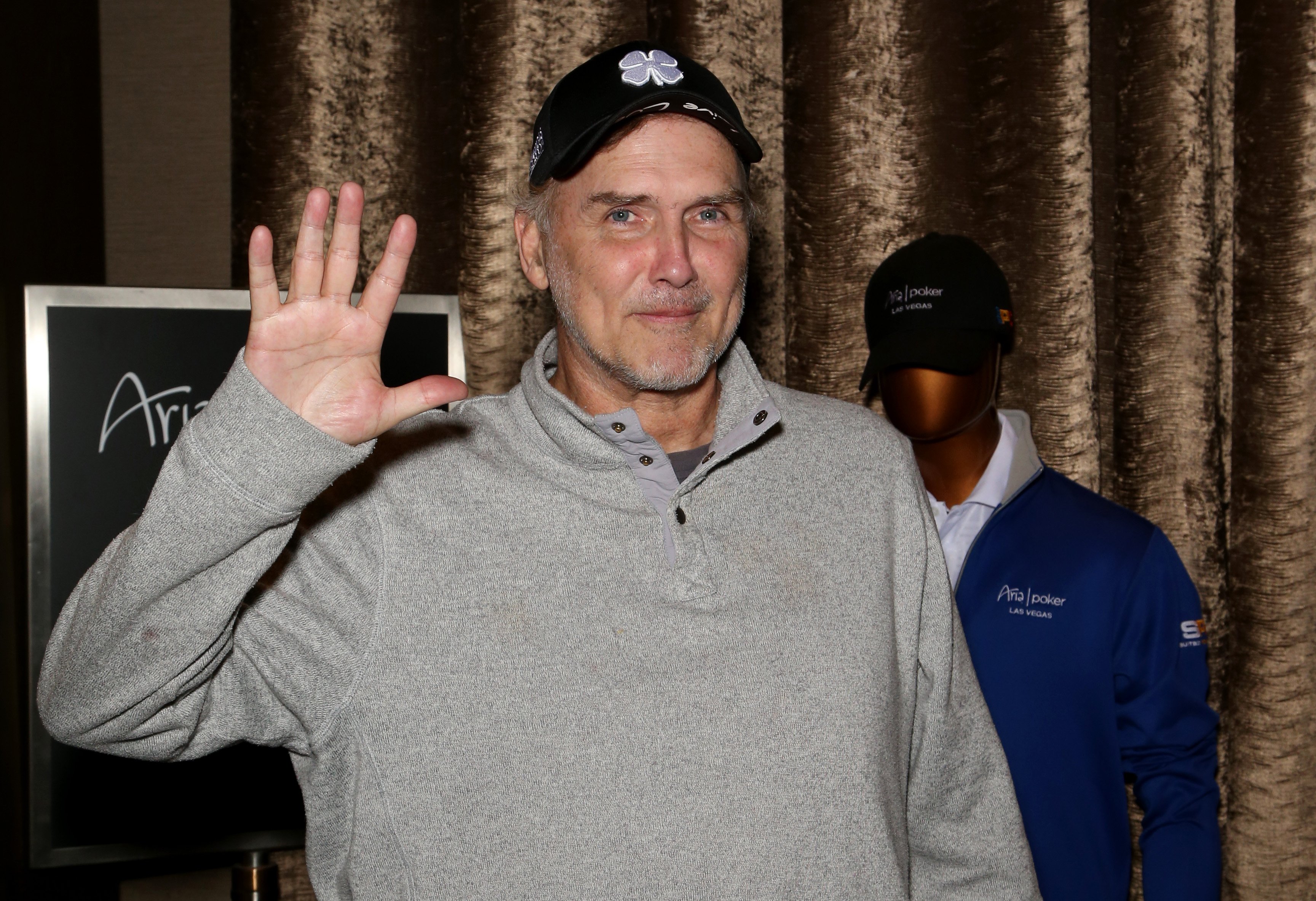 Norm Macdonald in a gray sweater and black baseball hat.