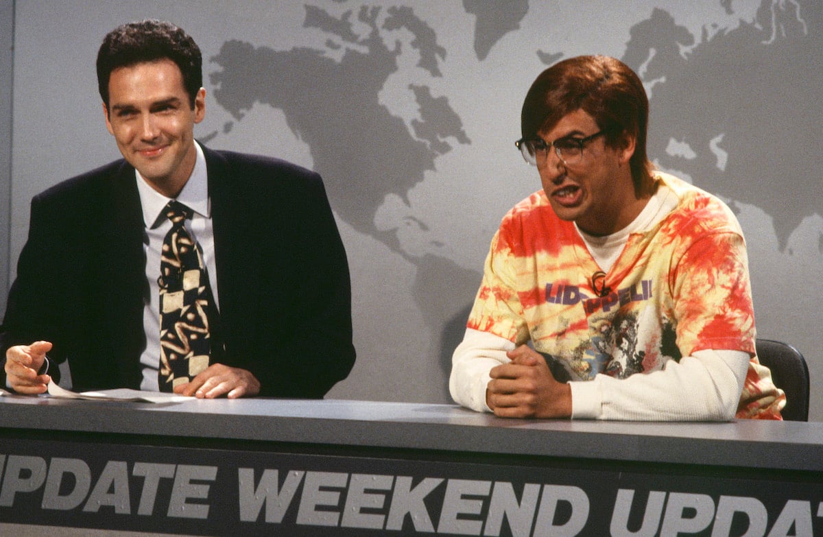 Norm Macdonald and Adam Sandler on 'Saturday Night Live' in 1994. They sit at the Weekend Update desk, Macdonald in a suit and Sandler in a bright orange shirt and black glasses and wig. In 1995, Macdonald and Sandler co-starred in 'Billy Madison,' their first film together.