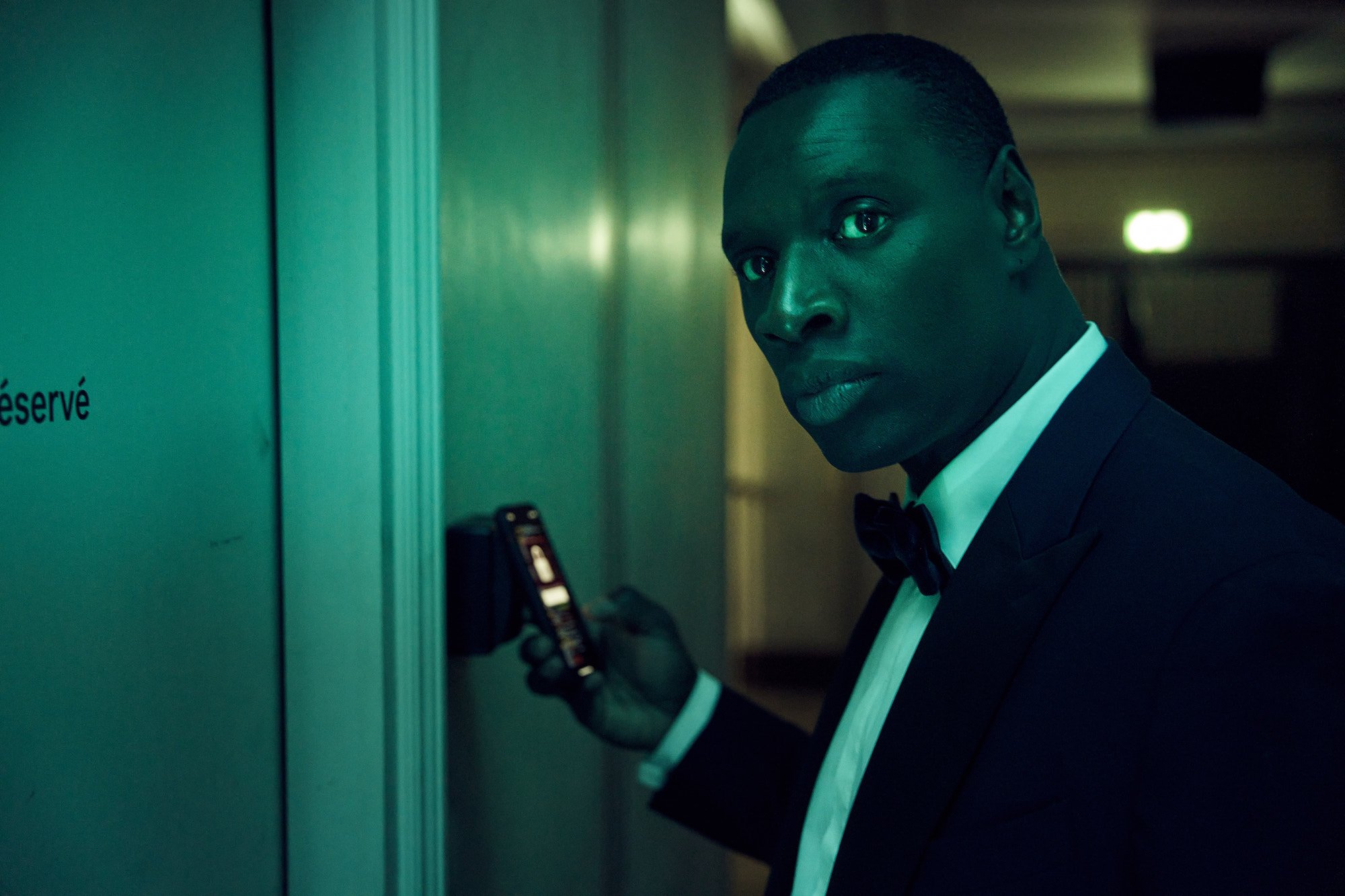 Omar Sy dressed in a black suit and holding a cellphone in 'Lupin.'