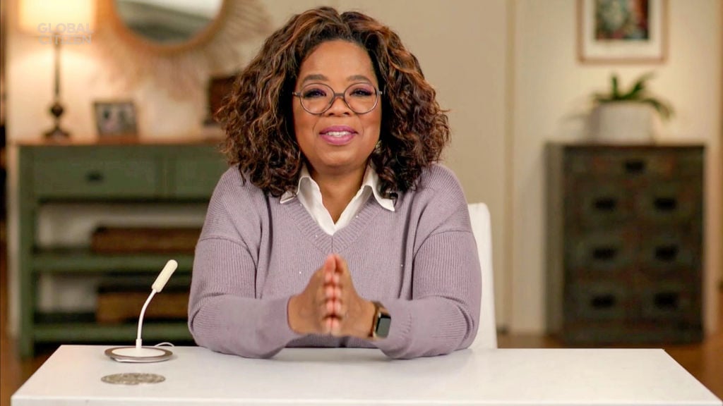 Oprah Winfrey speaks to the camera with her hands folded. She's wearing a lavender sweater and glasses.