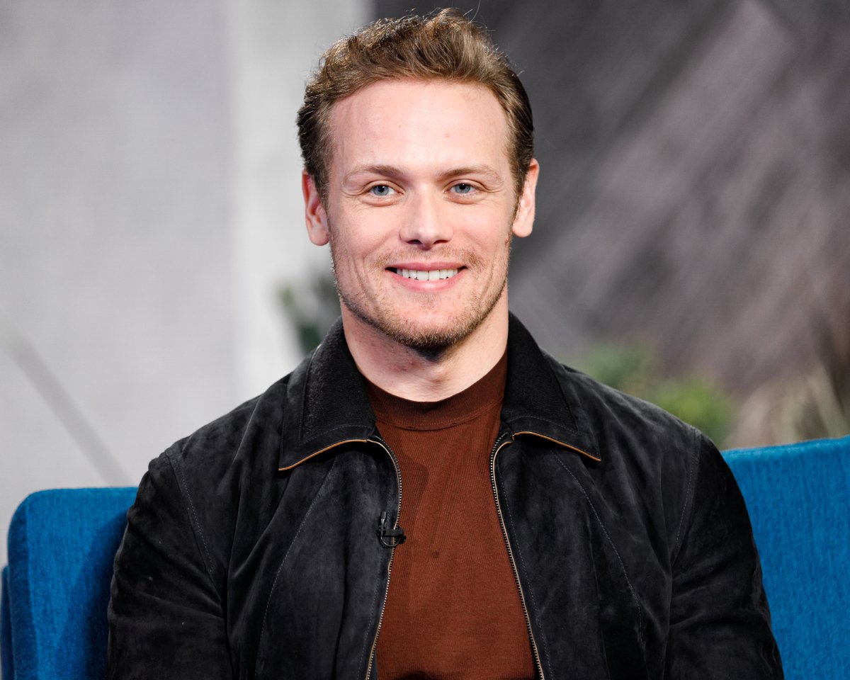 ‘Outlander’ star Sam Heughan visit’s 'The IMDb Show' on January 13, 2020 in Santa Monica, California. He wears a burnt orange shirt with a black suede jacket overtop and sits on a blue chair and smiles.