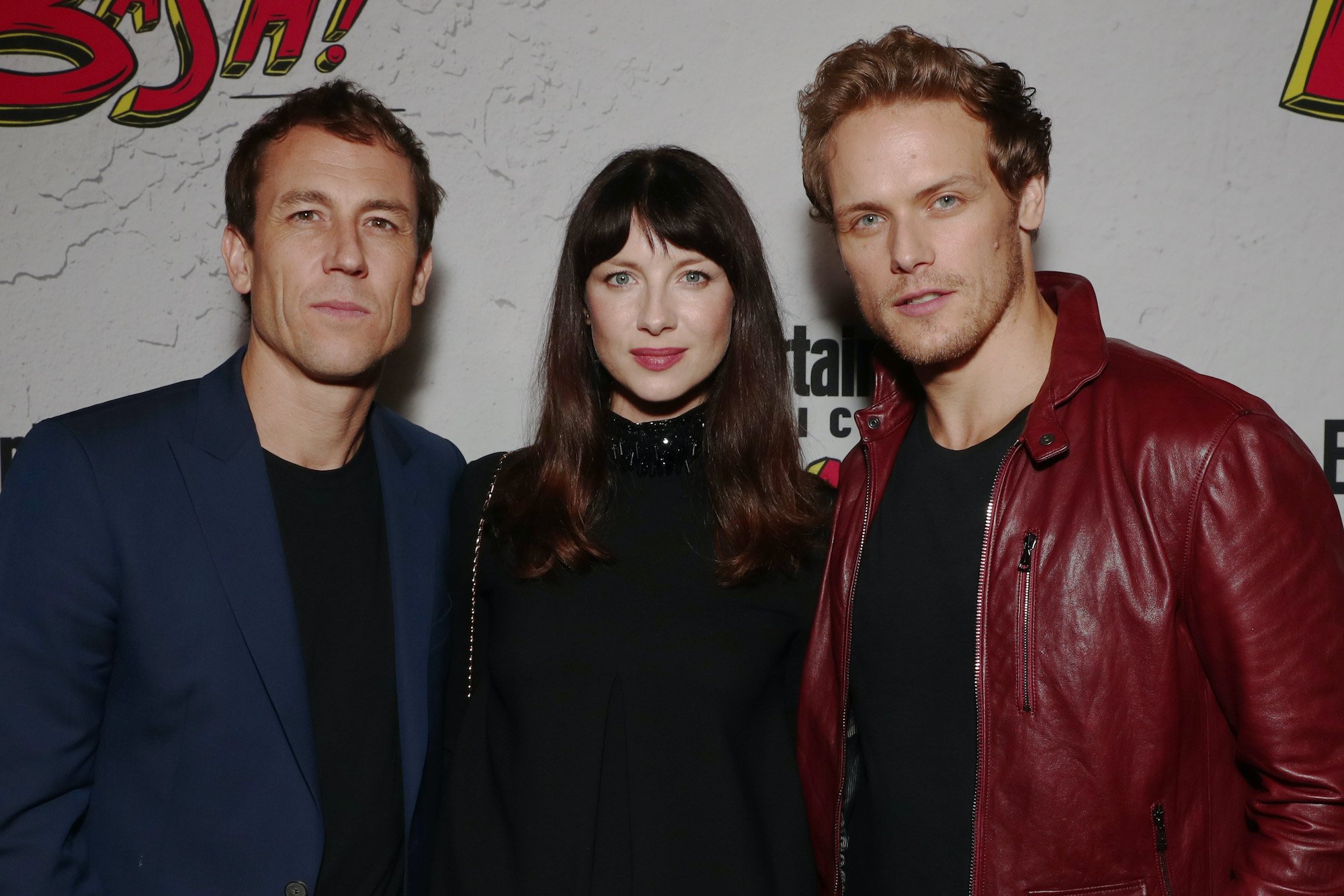 'Outlander' stars Tobias Menzies, Caitriona Balfe, and Sam Heughan pose at Comic-Con 2017 in San Diego in front of a white backdrop with red and yellow lettering. Menzies wears a black shirt and navy blue suit jacket. Balfe wears a black dress. Heughan wears a black shirt and red leather jacket. Heughan and Menzies shared many scenes in 'Outlander' as Jamie Fraser and Jack Randall. Menzies won an Emmy at the 2021 Emmy Awards on Sept. 19, and Heughan's reaction to Menzie's Emmy win was so supportive.