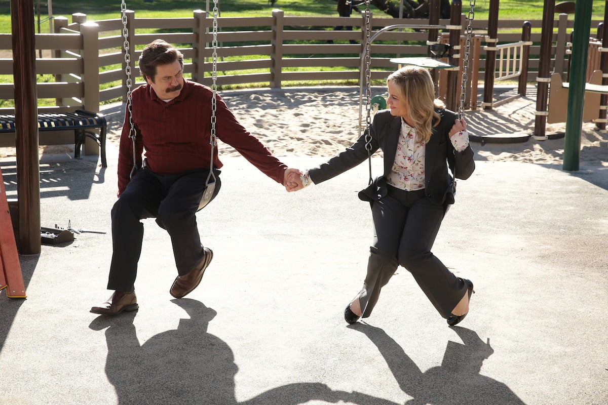 Ron Swanson and Lesli Knope holding hands on swings in an episode of 'Parks and Recreation'