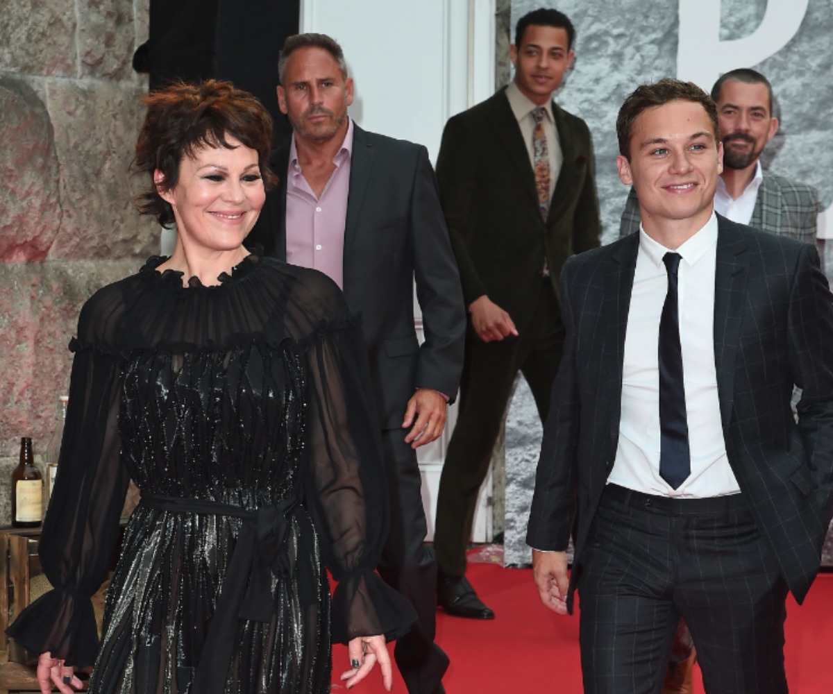Helen McCrory and Finn Cole walk beside each other and smile at the premiere of the 5th season of 'Peaky Blinders.' McCrory is wearing a long-sleeved black dress and Cole is wearing a suit and tie.
