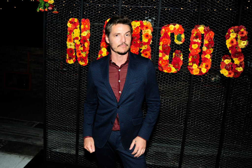 Pedro Pascal, dressed in a maroon shirt and navy jacket, poses in front of a 'Narcos' sign at a red carpet screening for the series.