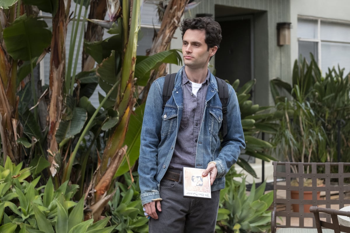 Joe Goldberg stands in front of his apartment in Los Angeles holding a book. 