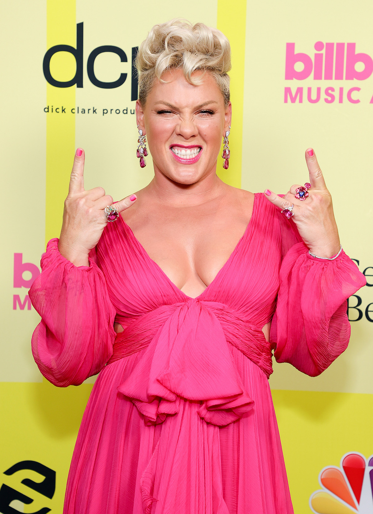 Pink wears a vibrant pink dress and short blonde hair, making rock signs with both hands and grimacing at the camera.