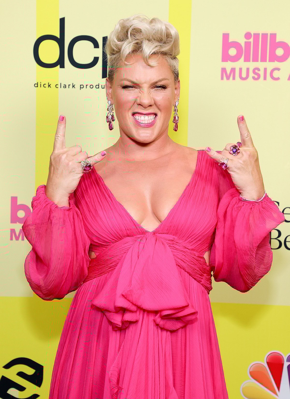 Pink wears a vibrant pink dress and short blonde hair, making rock signs with both hands and grimacing at the camera.