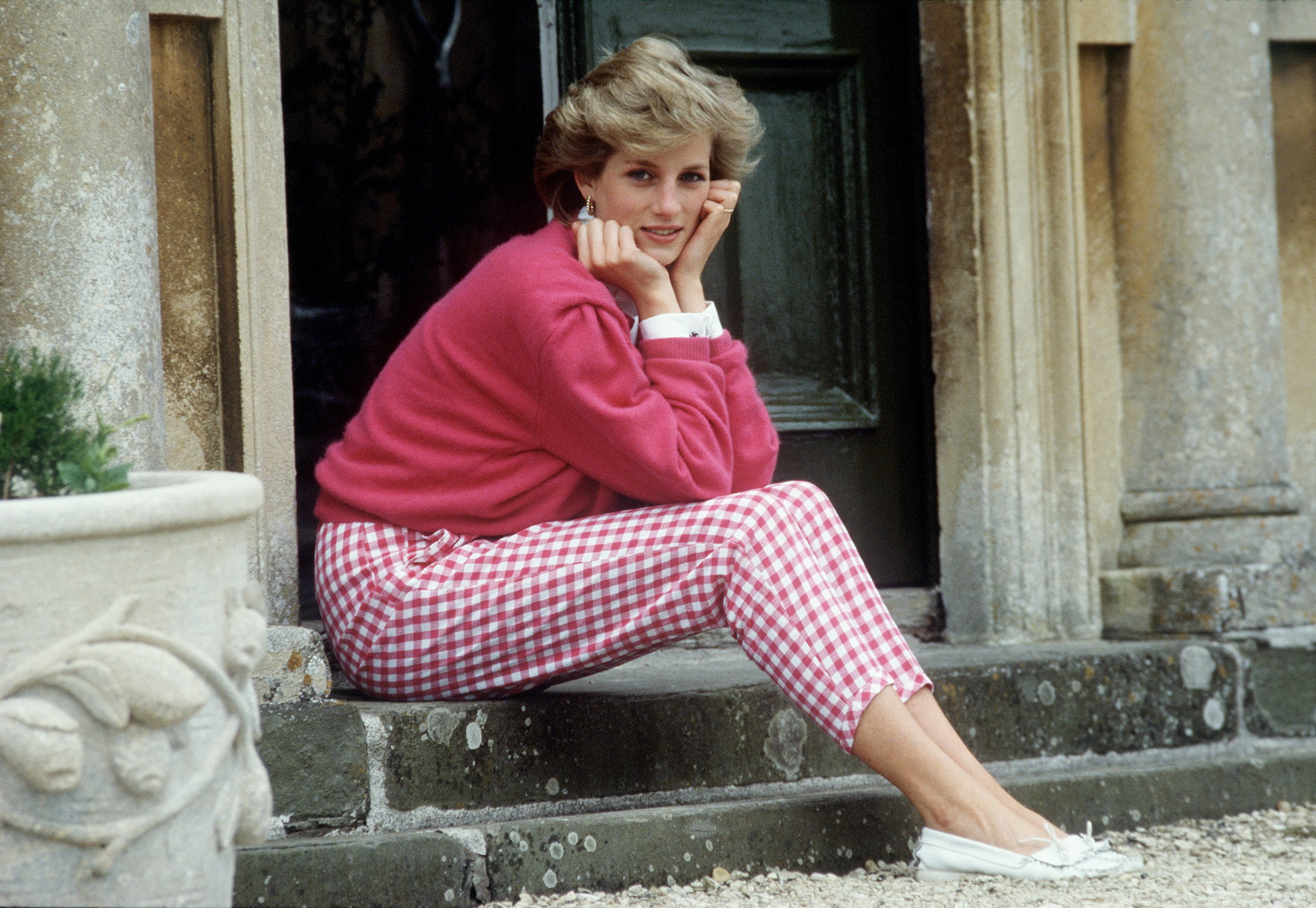 Princess Diana sitting on a step in a pink sweater and matching checkered pants