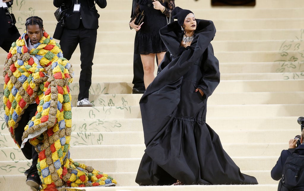 Rihanna and A$AP Rocky walk the white carpet for the MET Gala. Rihanna wears a long, black dress and Asap Rocky is covered in a colorful quit blanket.