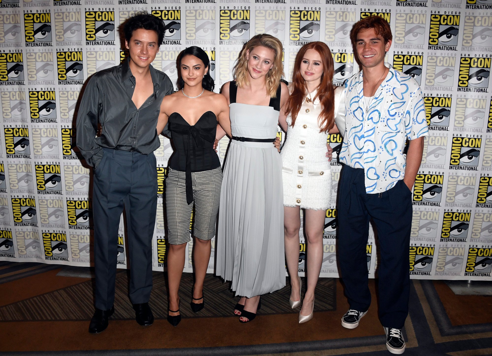 'Riverdale' Season 5 stars Cole Sprouse, Camila Mendes, Lili Reinhart, Madelaine Petsch, and K.J. Apa. They're standing side by side in front of a Comic-Con wall and wearing clothes that mix casual and formal styles.