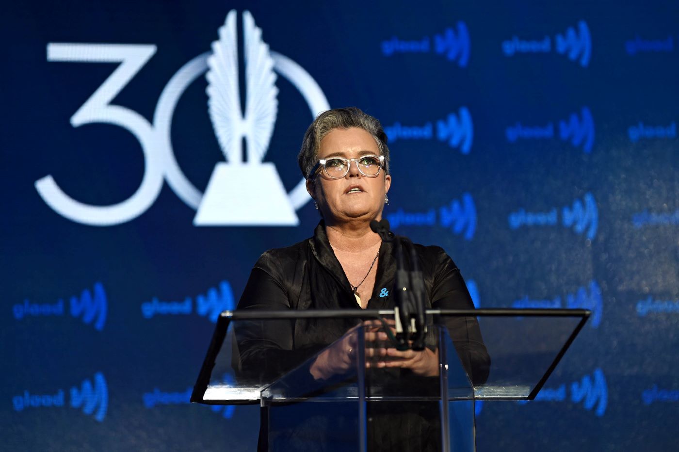 Rosie O'Donnell wearing all black in front of a blue and white background.