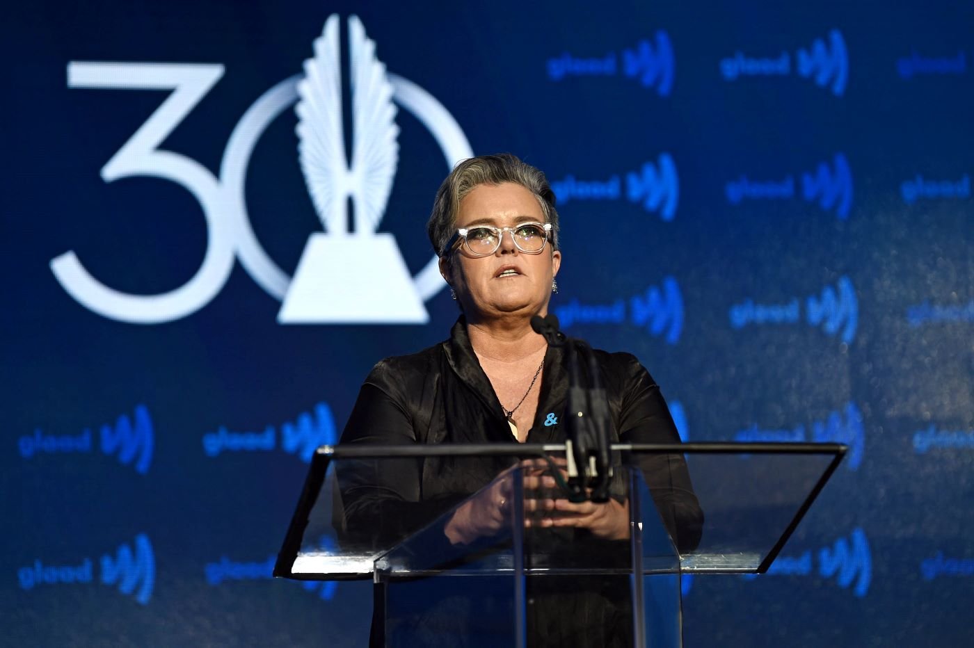 Rosie O'Donnell wearing all black in front of a blue and white background.