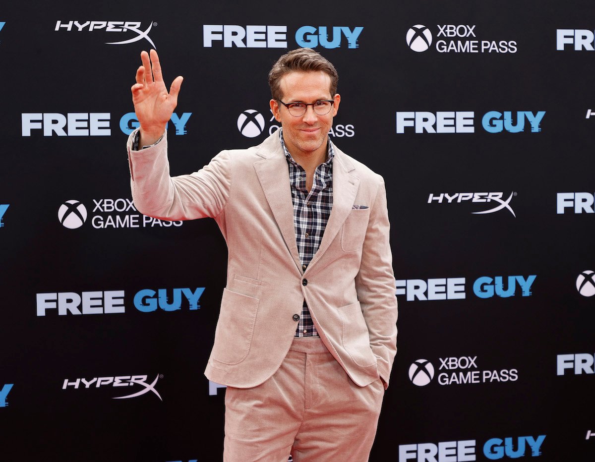 Ryan Reynolds wears a suit and glasses, waving for the camera at an event.