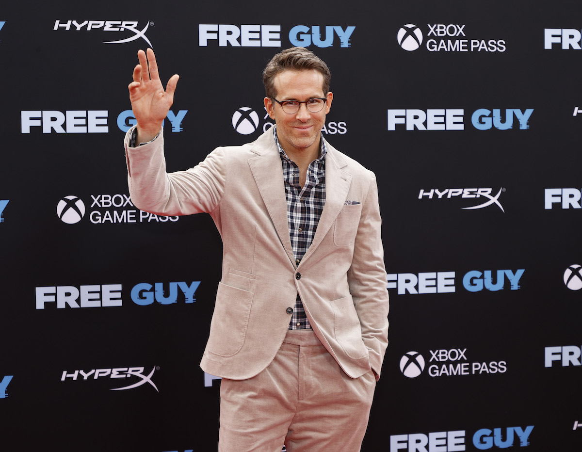 Ryan Reynolds wears a suit and glasses, waving for the camera at an event.