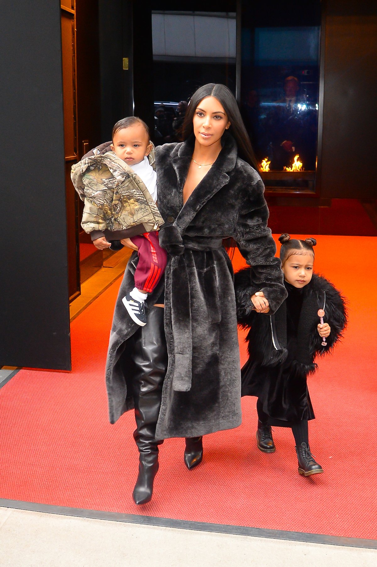 Kim Kardashian, wearing a long black coat, holds son Saint West in one arm while guiding daughter North West with the other.