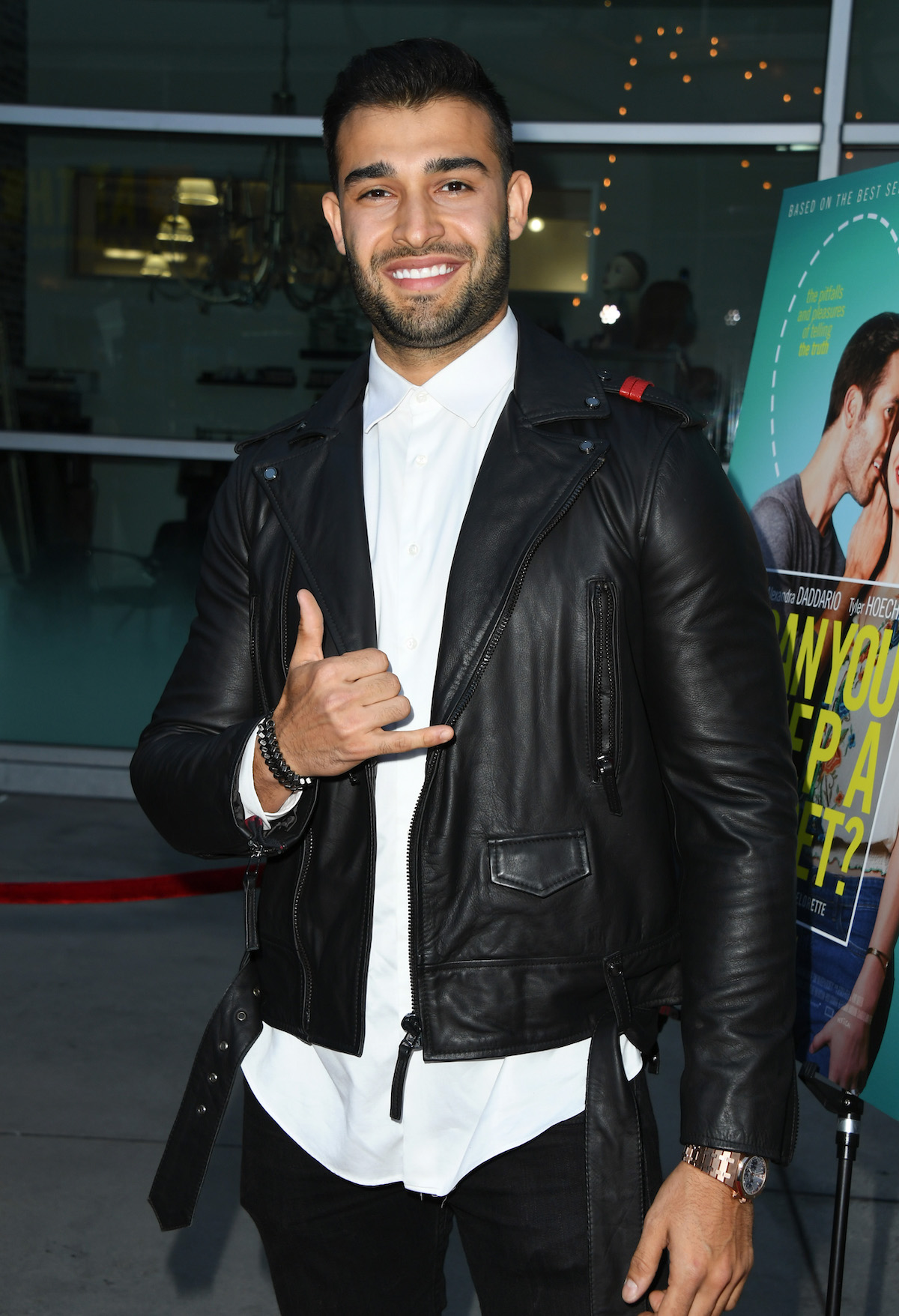 Sam Asghari flashes the hang loose sign with one hand while wearing a black leather jacket and smiling.