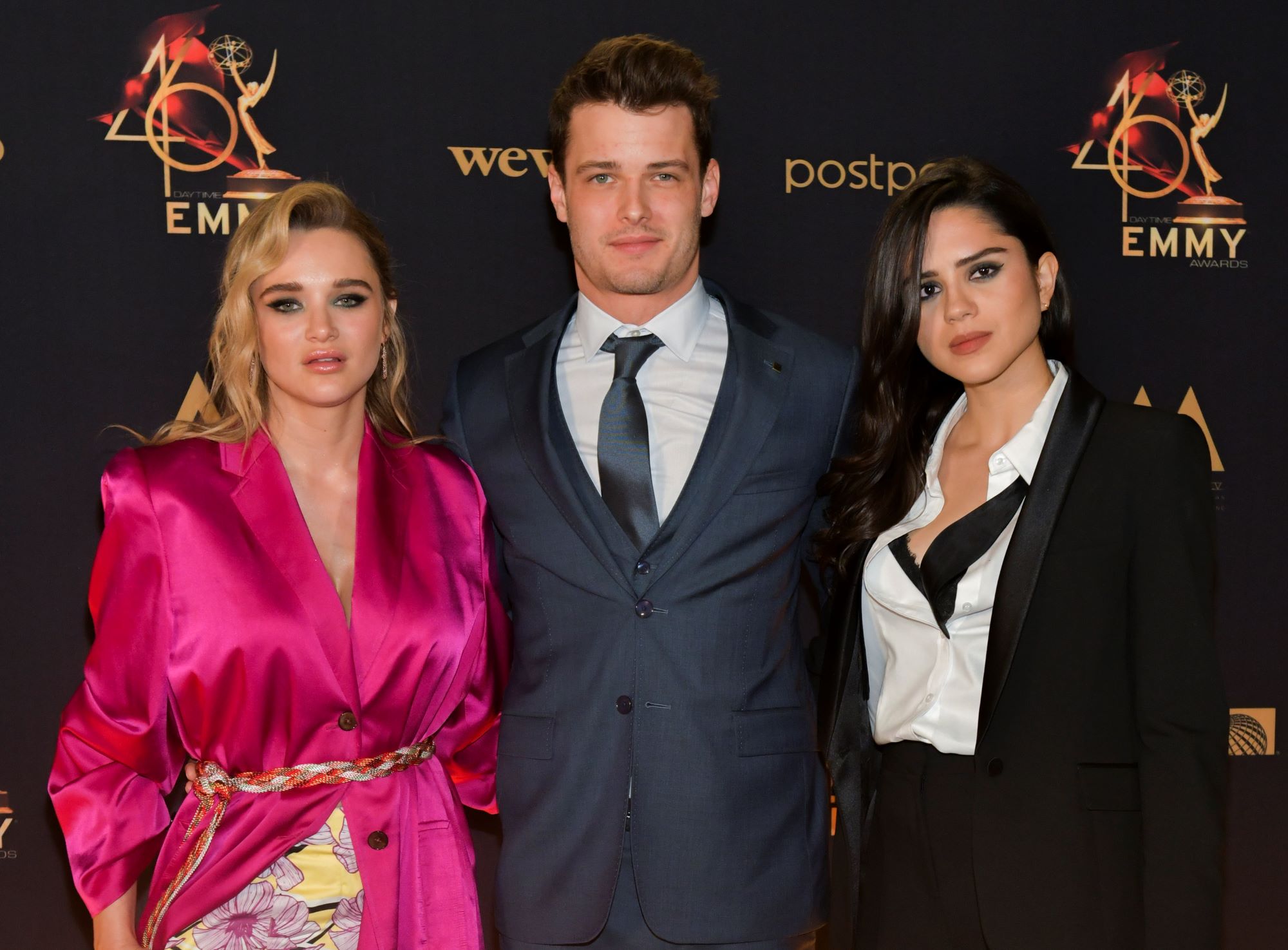 Actors Hunter King, Michael Mealor, and Sasha Calle huddle together and pose for a picture on the red carpet. Hunter wears a pink suit jacket, Michael wears a dark suit and tie, and Sasha wears a black suit over a white button up shirt.