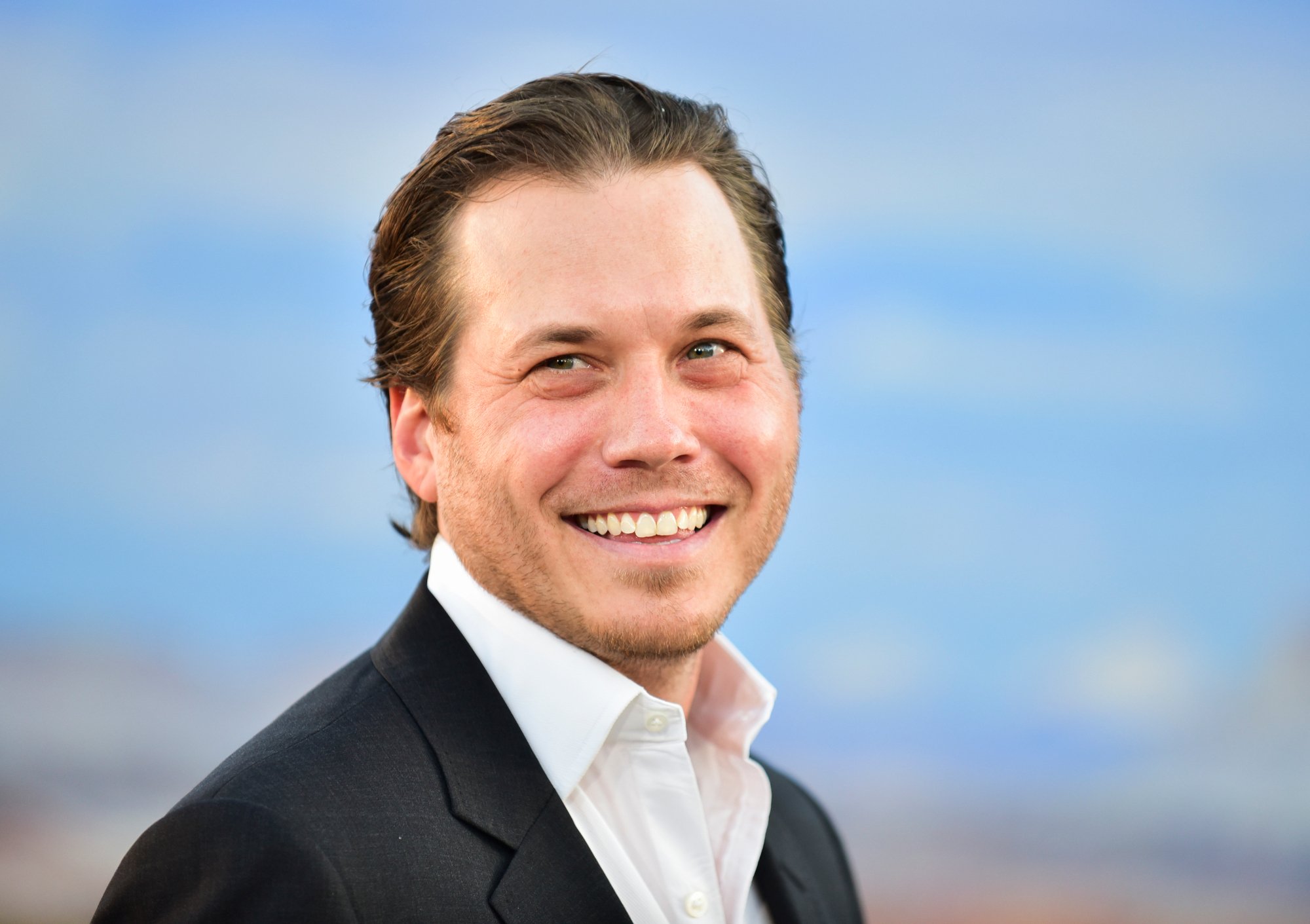 Scott MacArthur, who plays Adam in 'Lucifer' Season 6. He's wearing a white, collared shirt and black jacket and smiling. The blurred background behind him is blue.